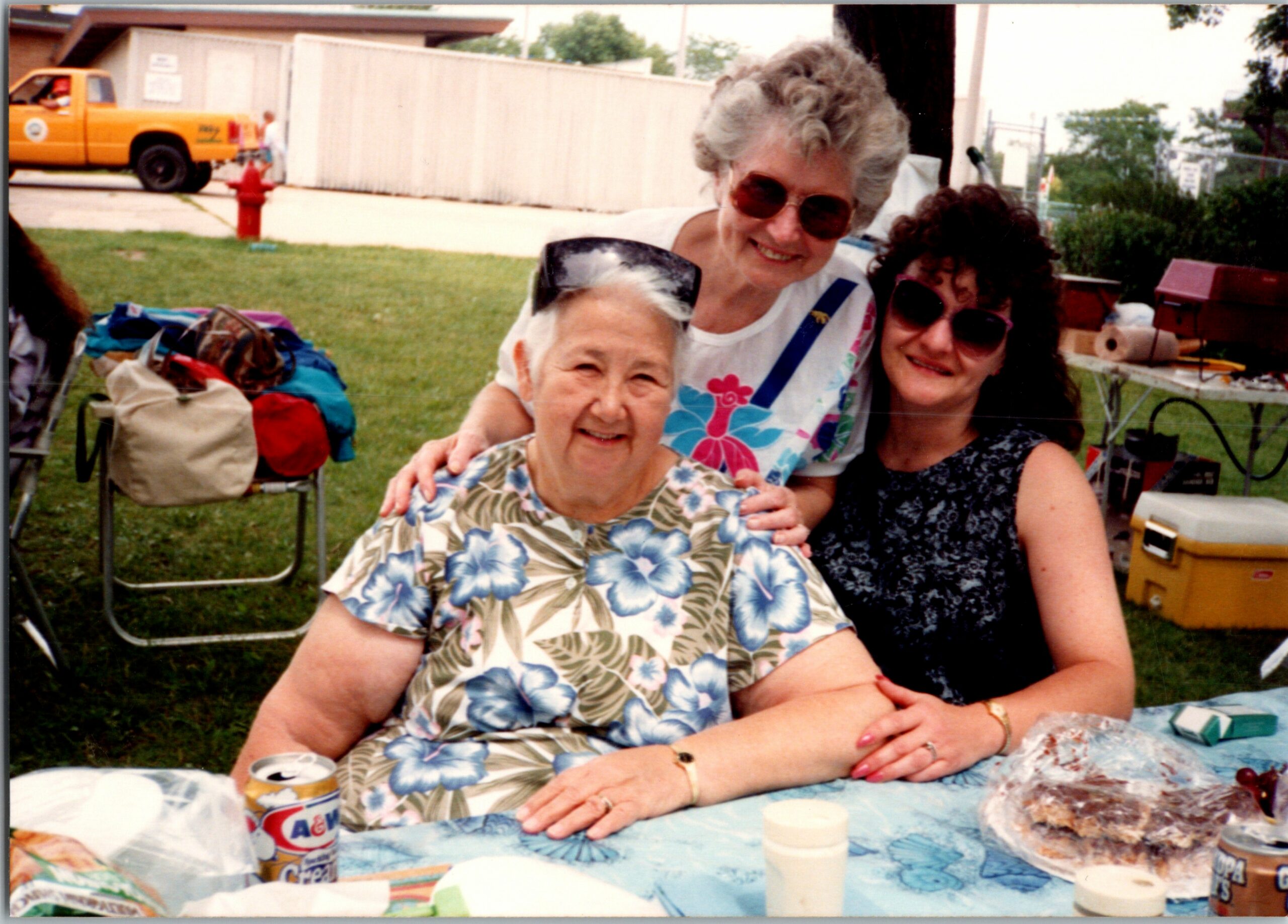 Three women smiling at an outdoor backyard picnic table; one in a floral shirt in front, a gray-haired woman with sunglasses behind, and a fourth person to the right; table has cans and food items.