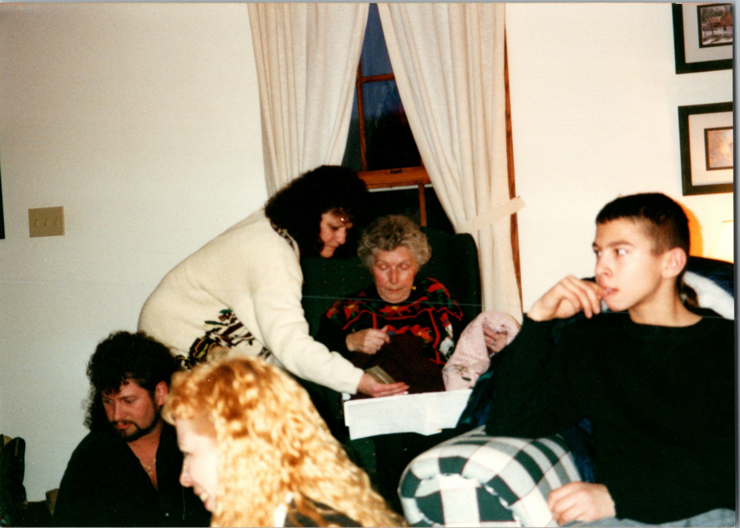 People gathered around a dining table in a living room; an older woman appears to be knitting or looking at something on the table while others chat/leisurely interact in the background.