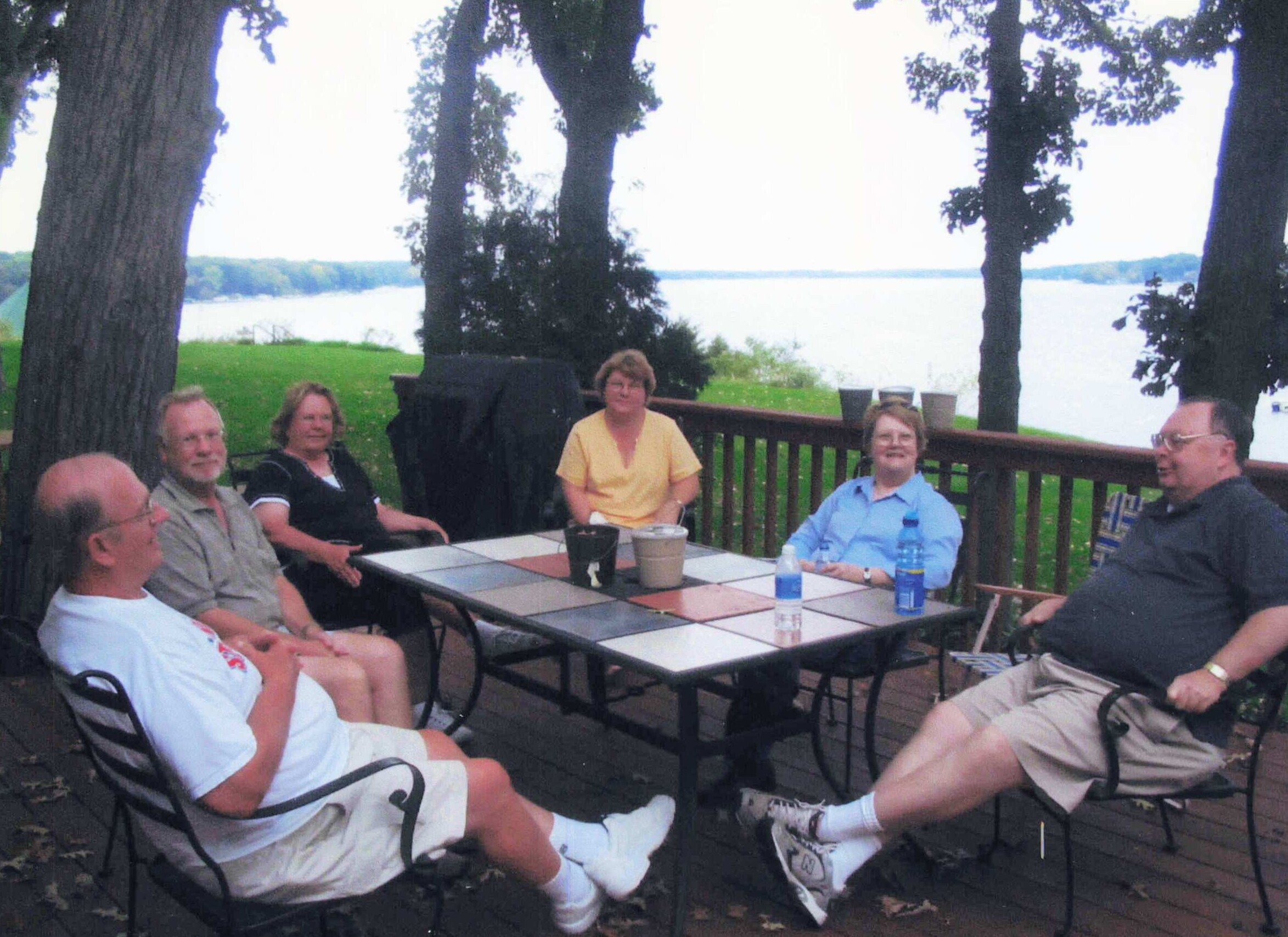 Group of adults sitting around a tiled outdoor table on a wooden deck by a lake, surrounded by trees. In the background is water and greenery.
