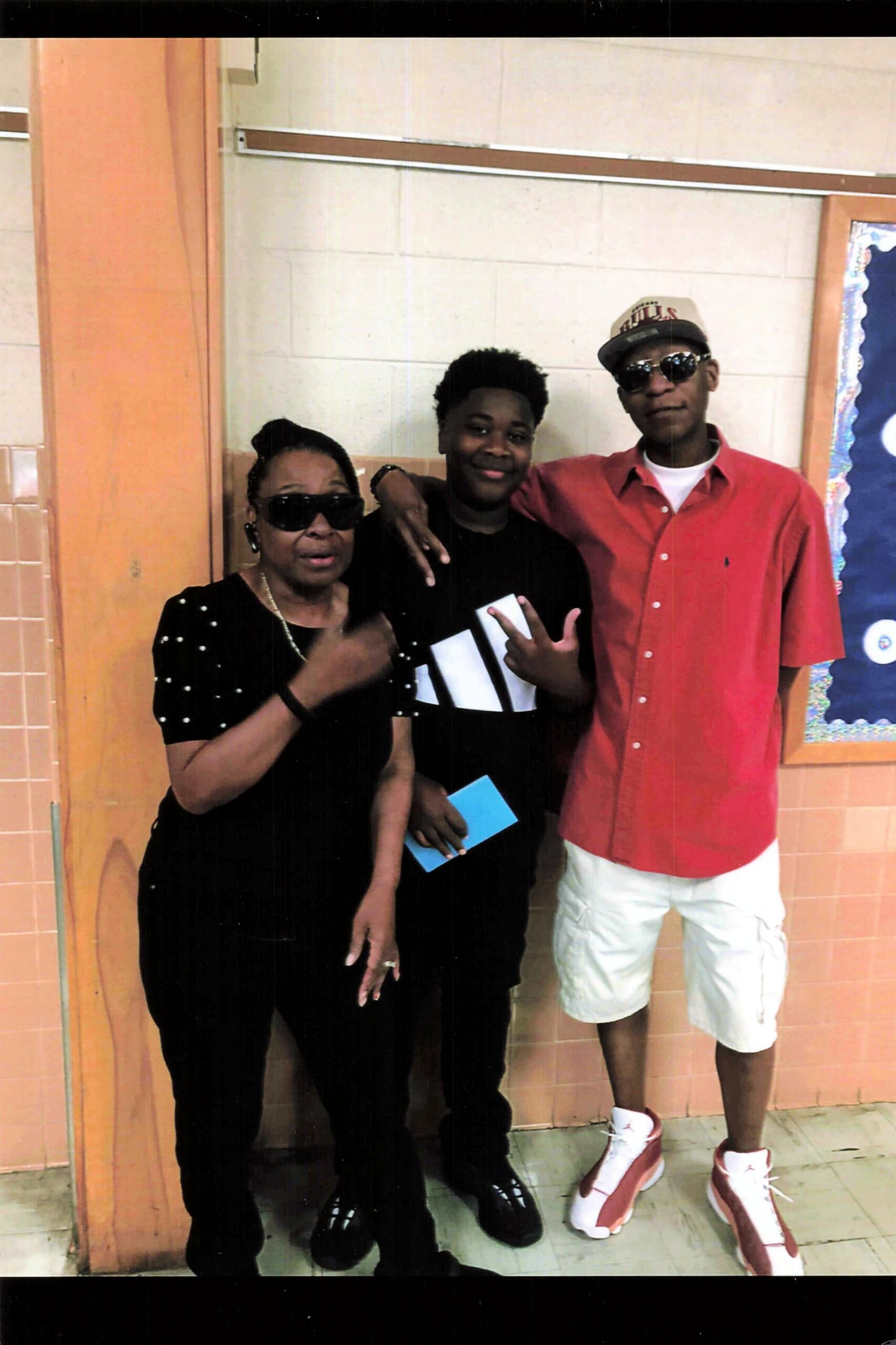 Three people posing in a school hallway: a woman in black with sunglasses on the left, a person in black center holding a blue card, and a man in a red shirt with a cap and sunglasses on the right.