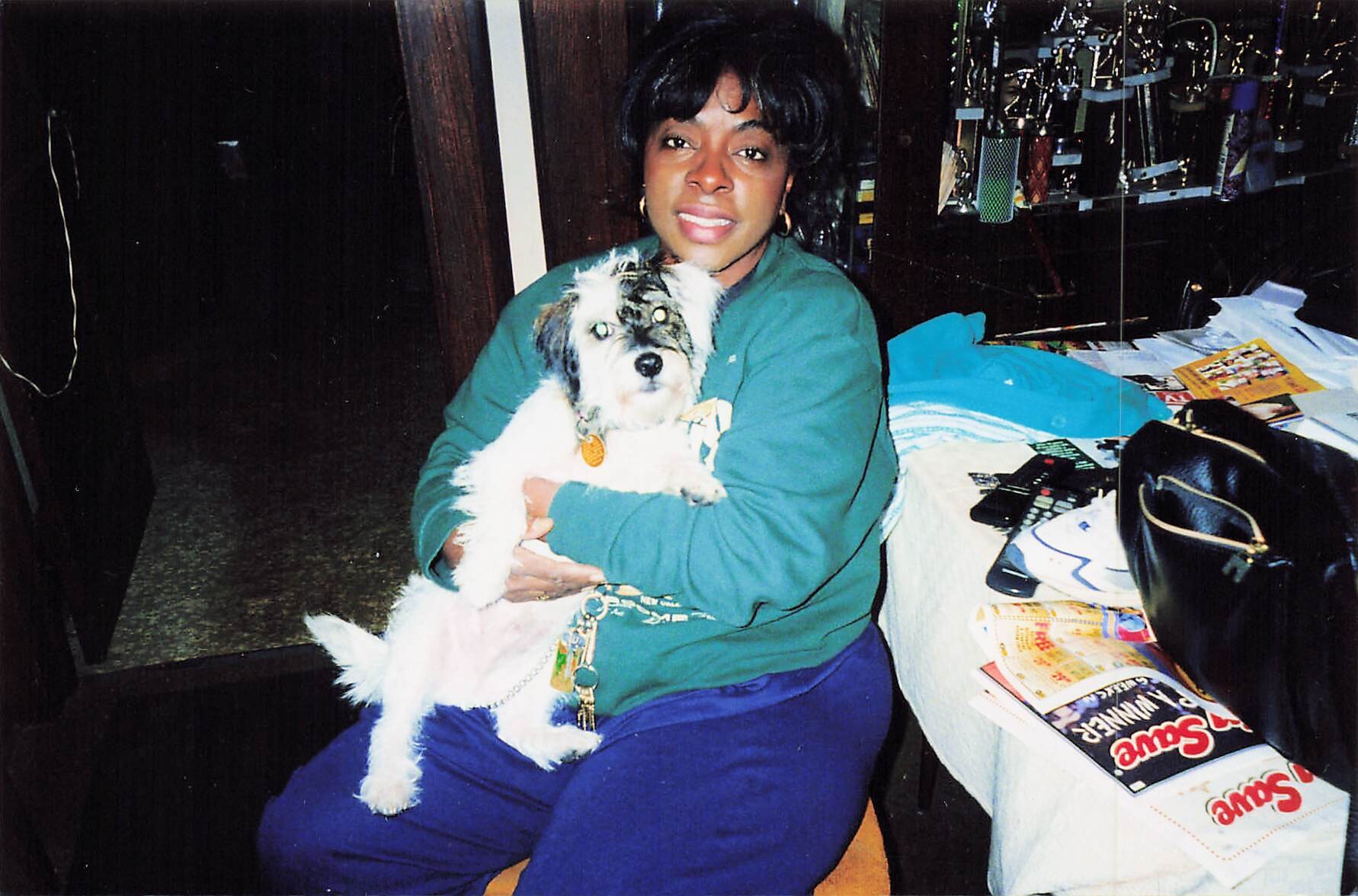 Woman in a teal sweatshirt sits on a chair, smiling while holding a fluffy white-and-gray dog in her lap indoors.