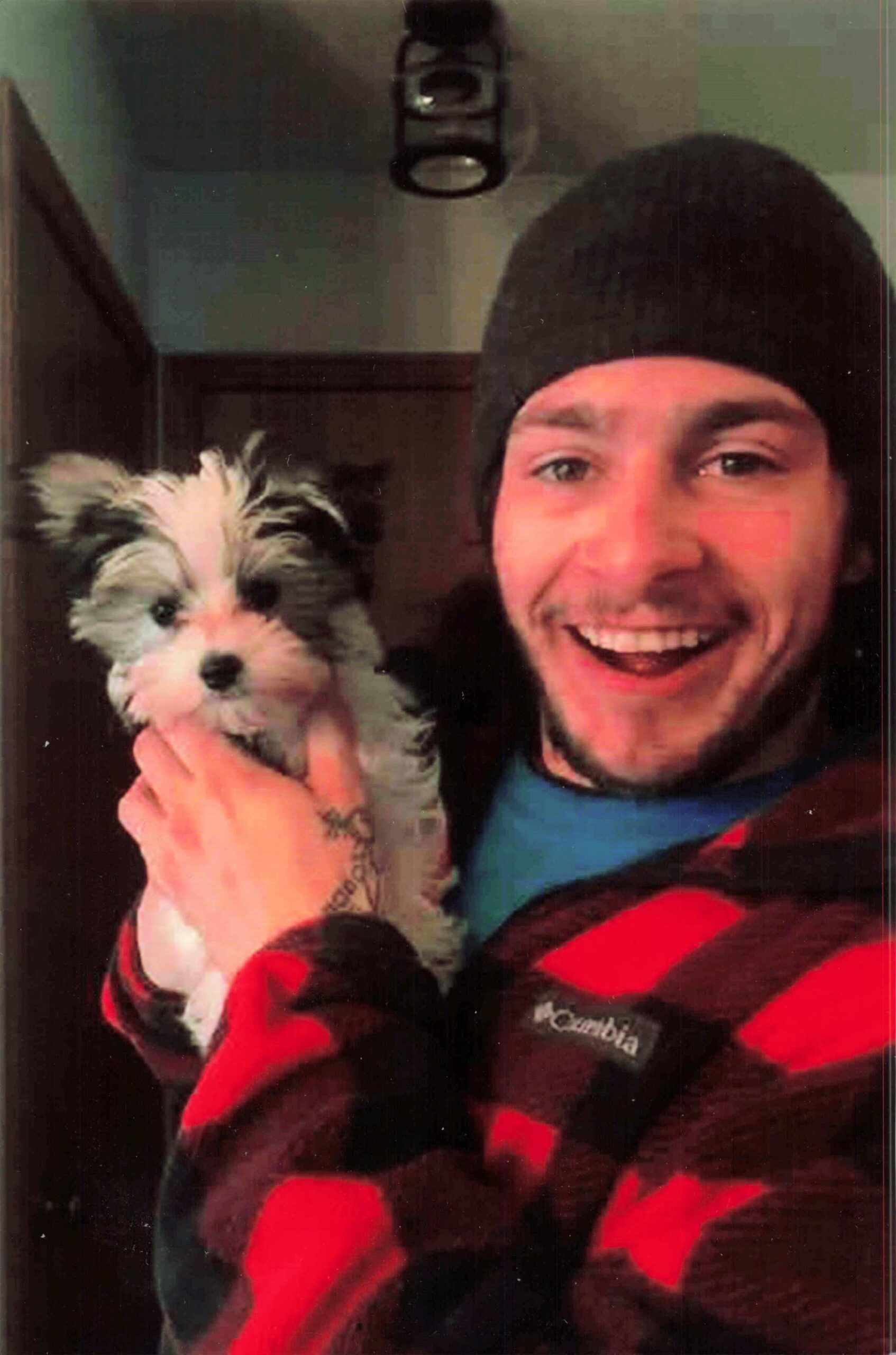 Smiling man in a black beanie holding a fluffy puppy indoors, both looking at the camera.