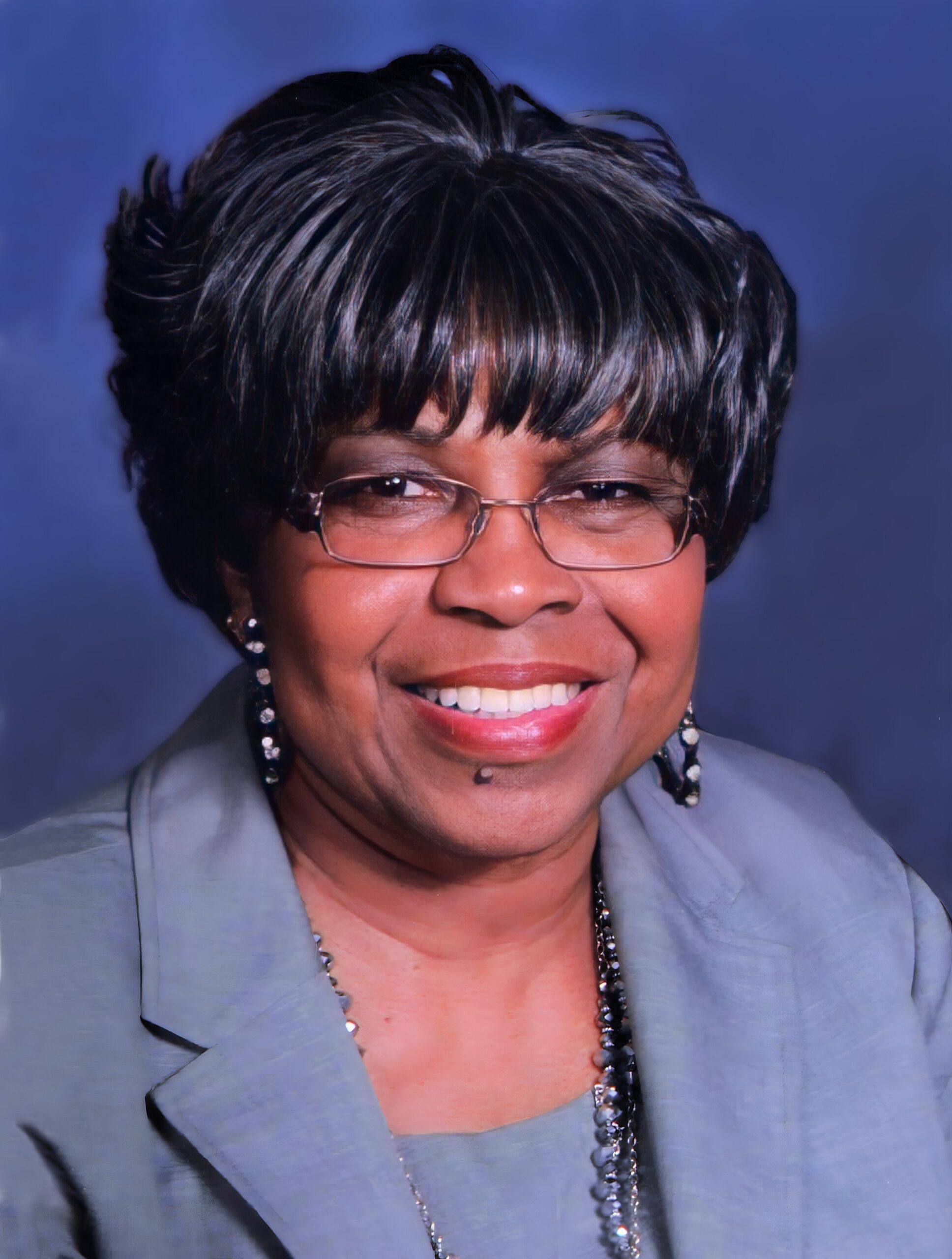 Smiling woman with short dark hair and glasses, wearing a light blue blazer against a blue background (close-up portrait).