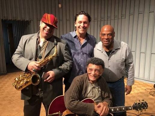 Four musicians pose together in a backstage rehearsal area; one man holds a saxophone, another sits with a guitar, and two others stand smiling beside him.