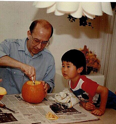 Older man carves a pumpkin at a table while a young boy watches closely, newspapers spread out before them.