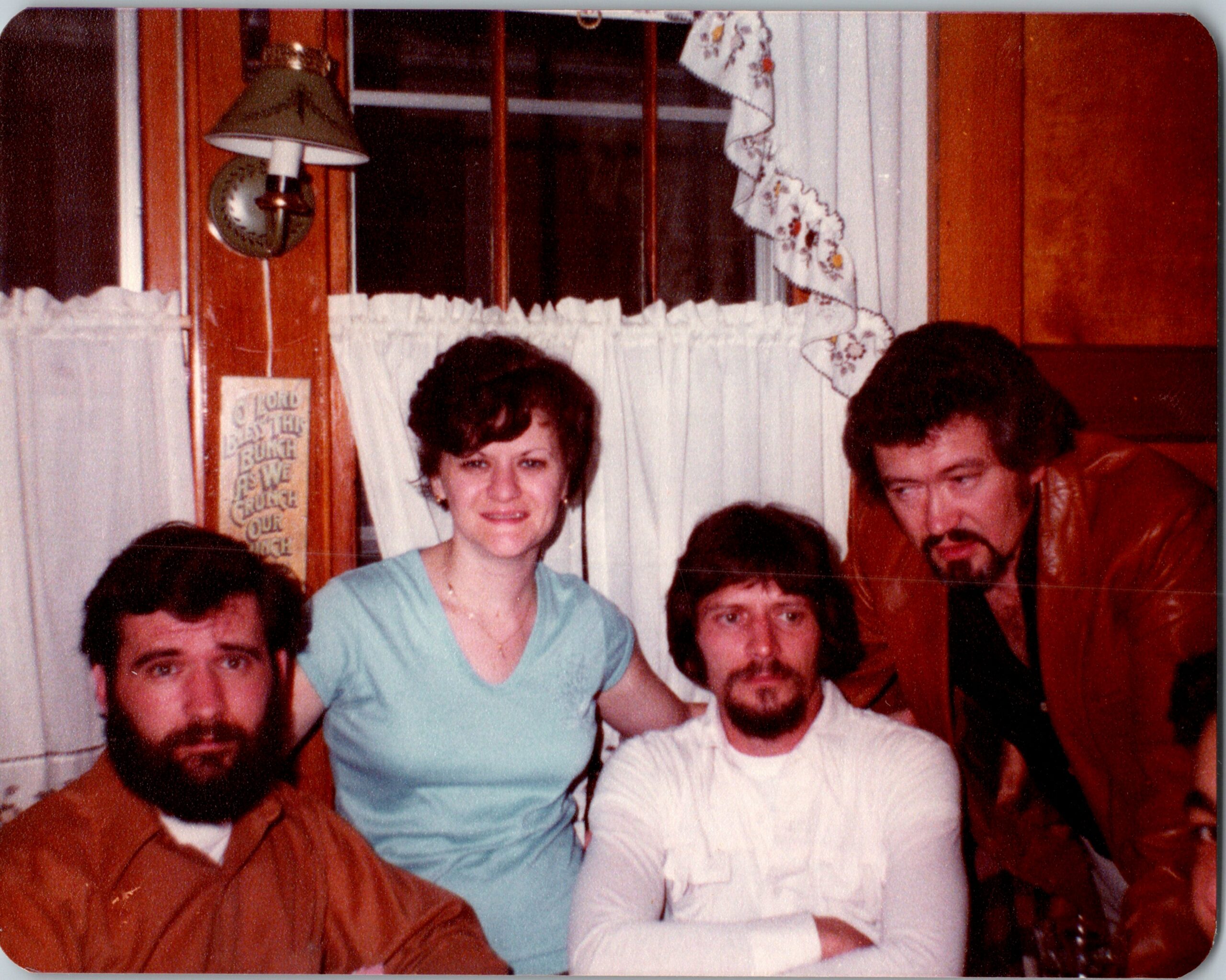 Group of adults posing for a casual photo in a wood-paneled room with lace curtains; woman in a blue dress sits center with men nearby.