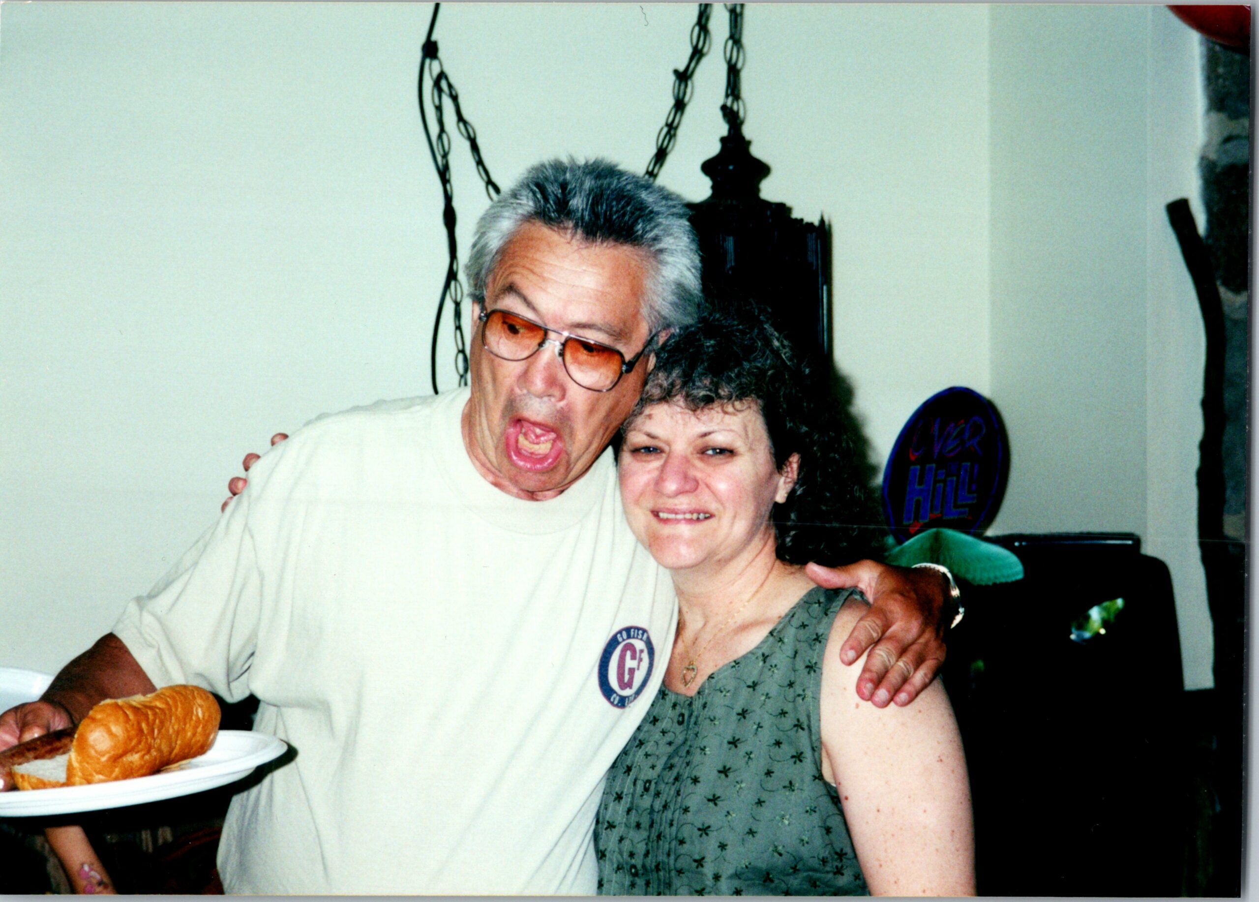 Smiling man with gray hair and orange-tinted glasses hugs a woman while holding a plate of bread at a casual gathering indoors.