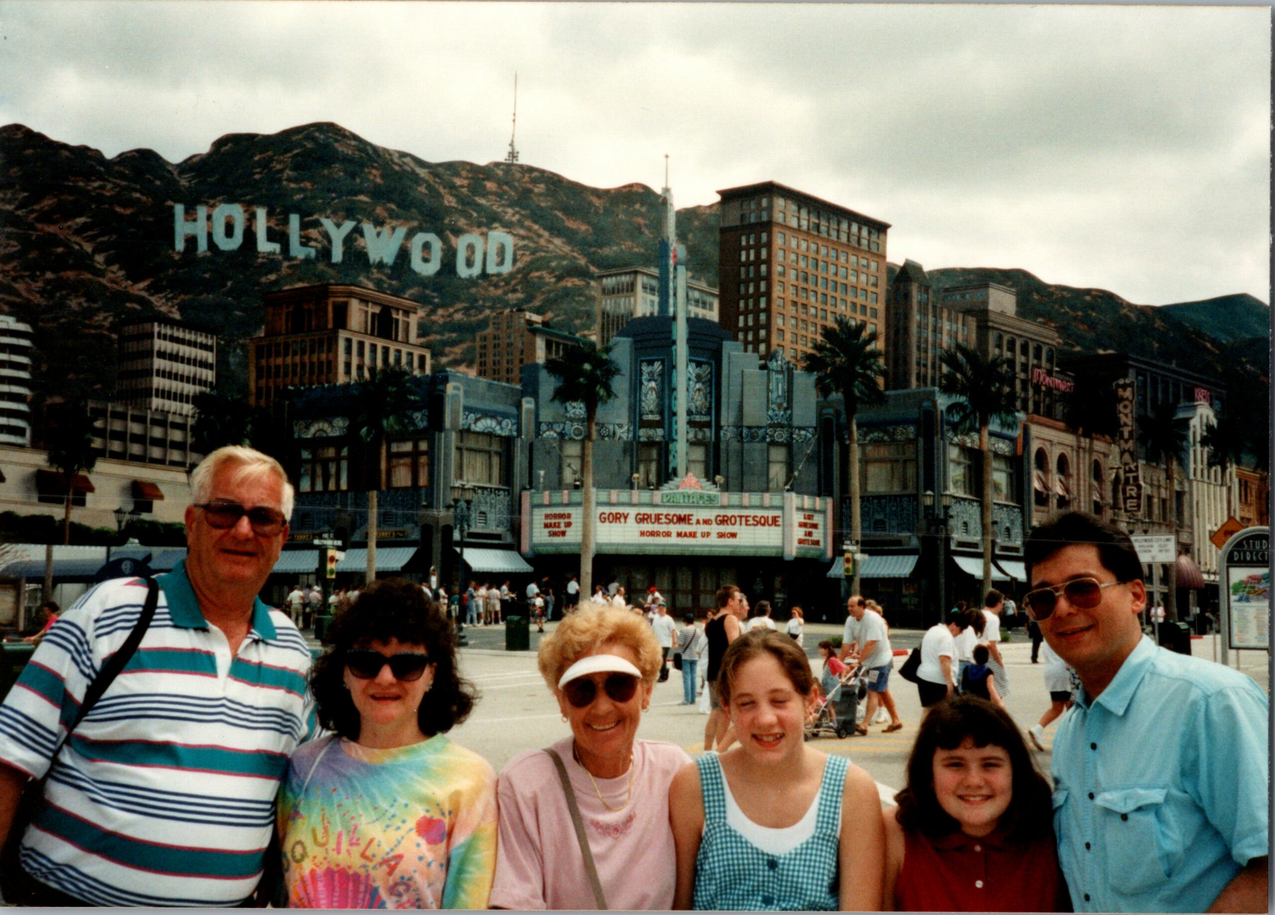 A group of smiling tourists posing for a photo in front of the Hollywood skyline with the iconic sign and themed buildings in the background.