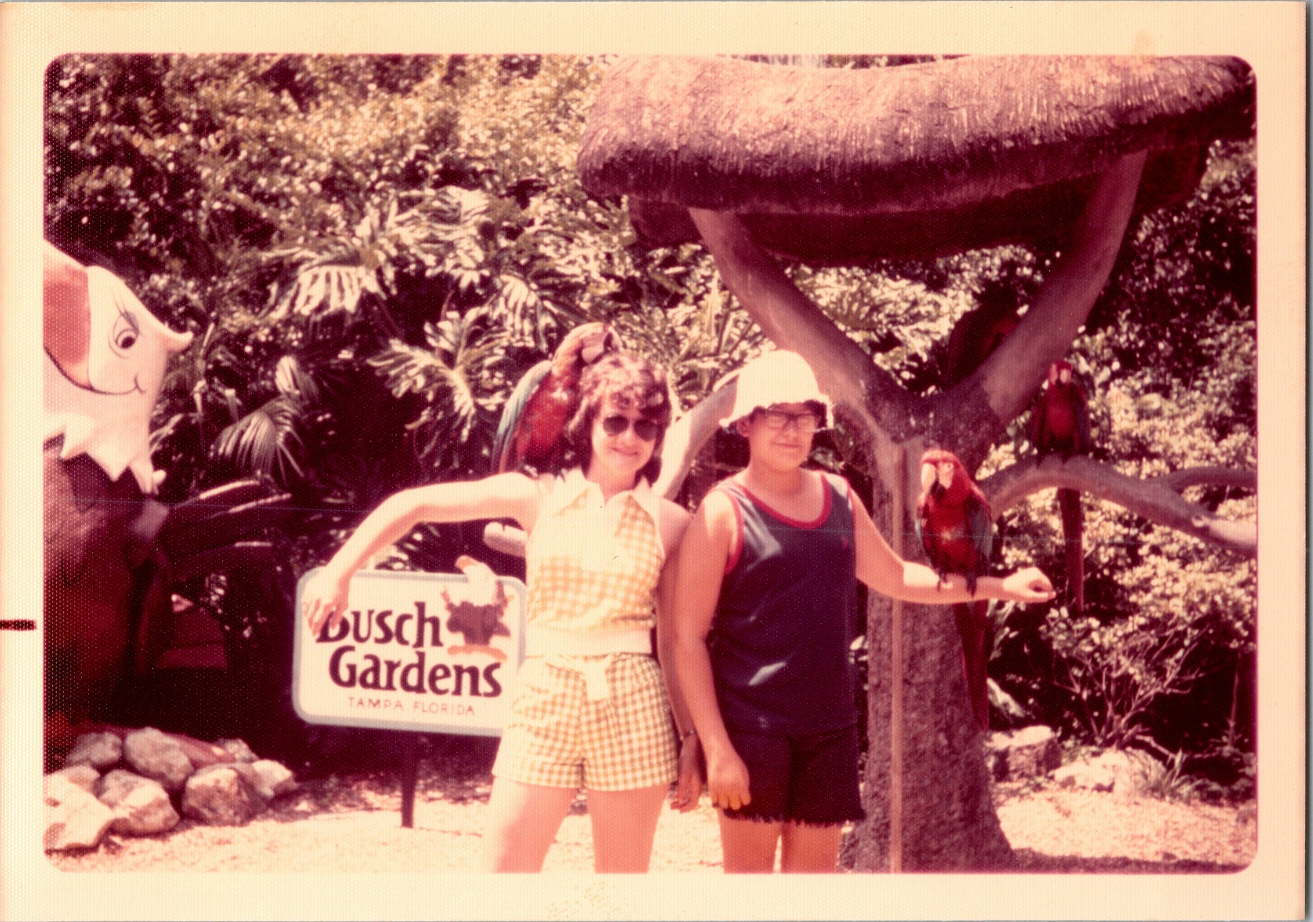 Two women pose in front of a Busch Gardens sign at Tampa, Florida, with tropical plants behind them.
