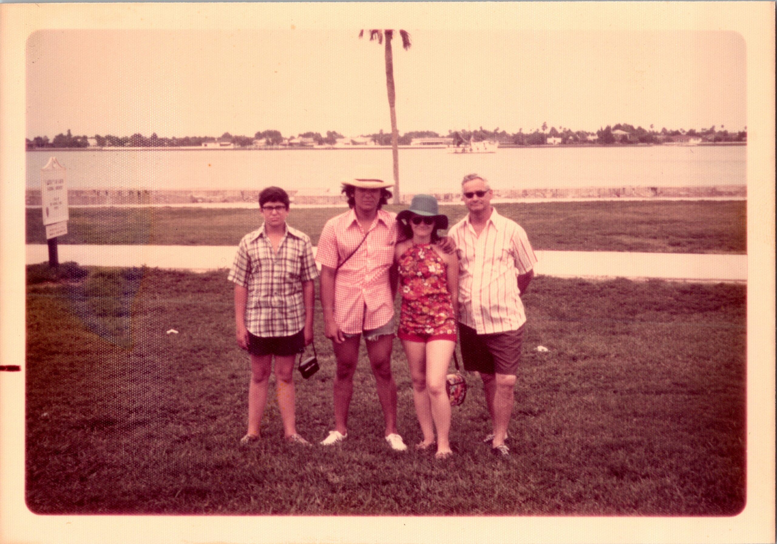 Four people pose together on a grassy lakeside, wearing summer clothing and hats, with water and distant houses in the background.