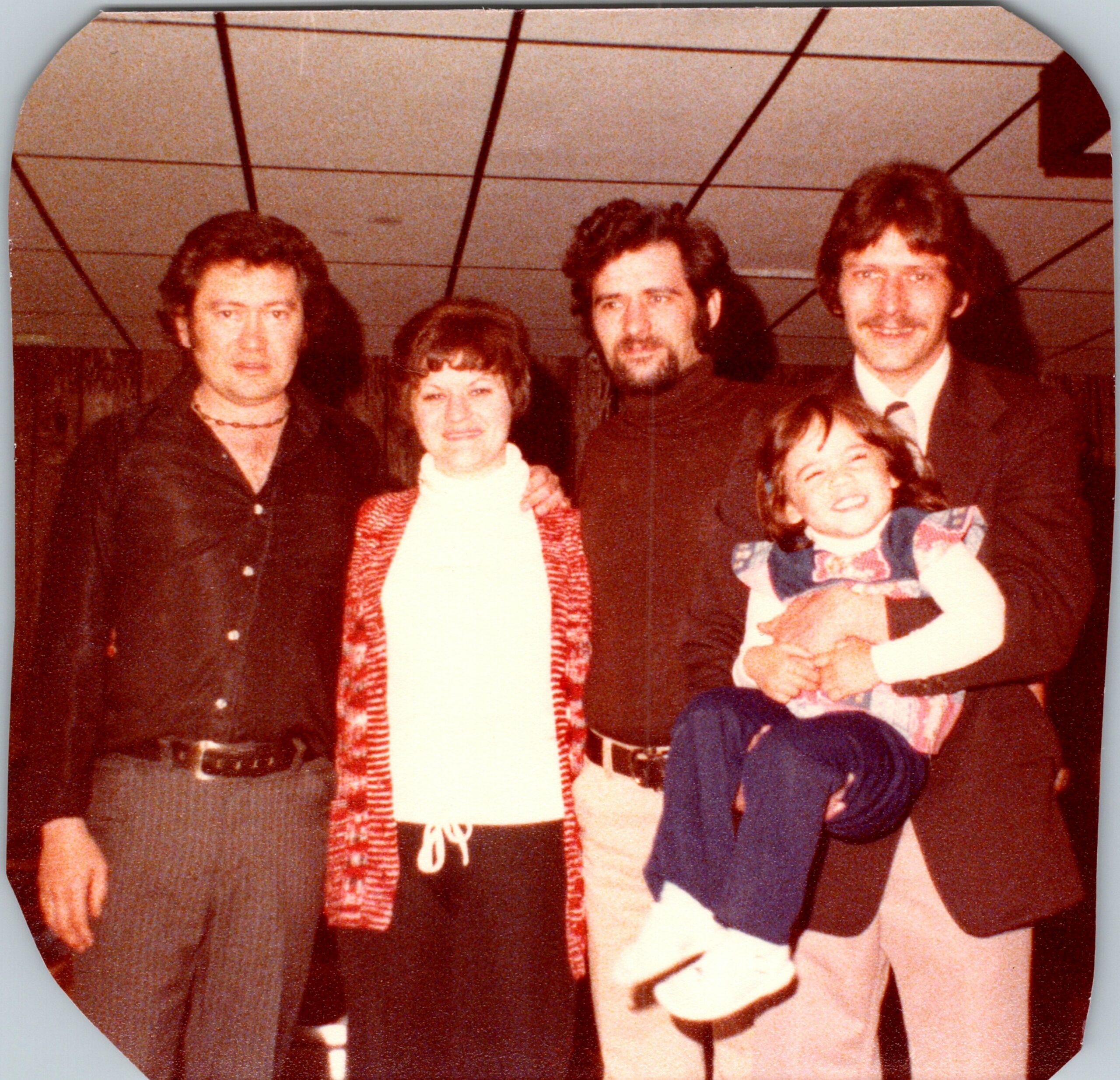 Group of adults posing together for a warm, indoor, social photo; several are smiling and wearing 1980s-style clothing.