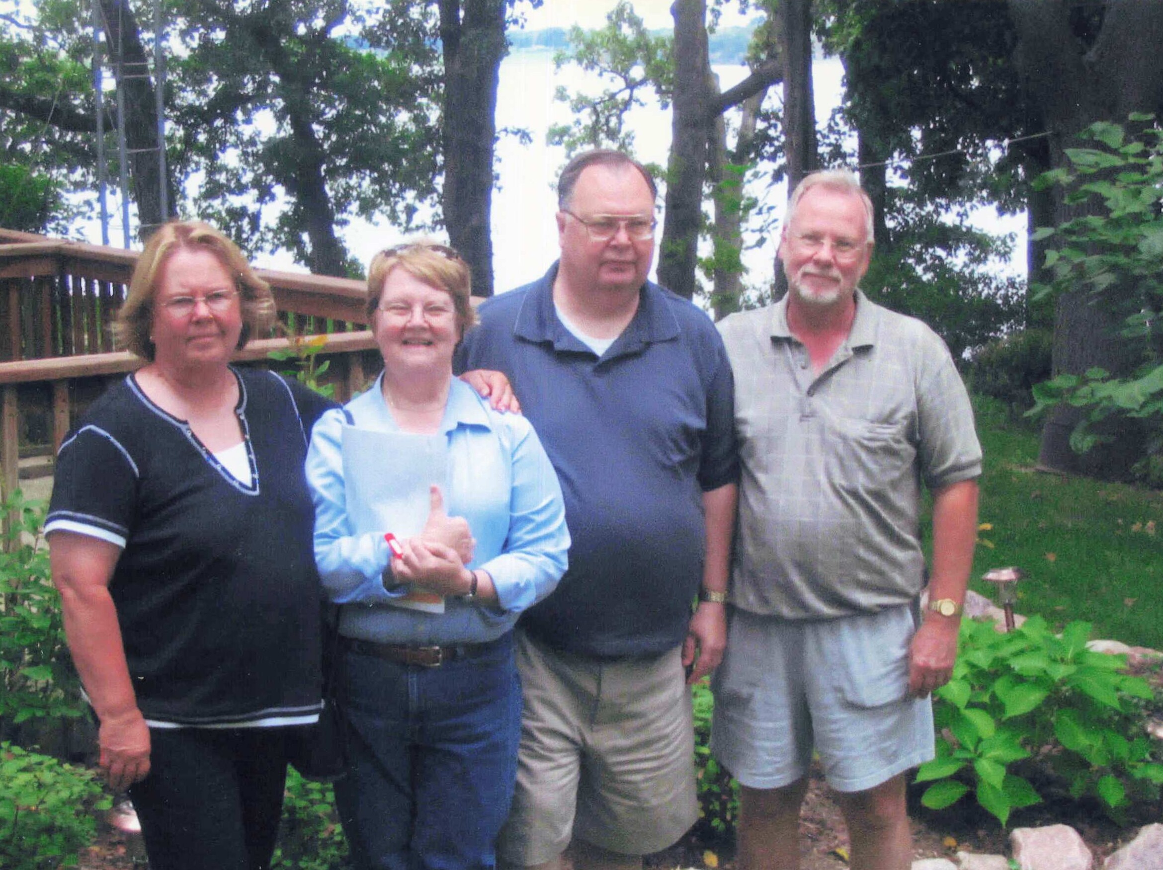 Group of four adults standing together outdoors, smiling, with trees and a wooden fence behind them in a garden setting.