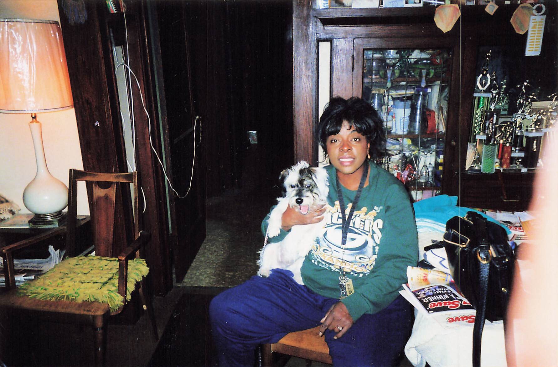 Woman sitting on a chair indoors, holding a small fluffy dog, in a room with a tall cabinet and trophies in the background.