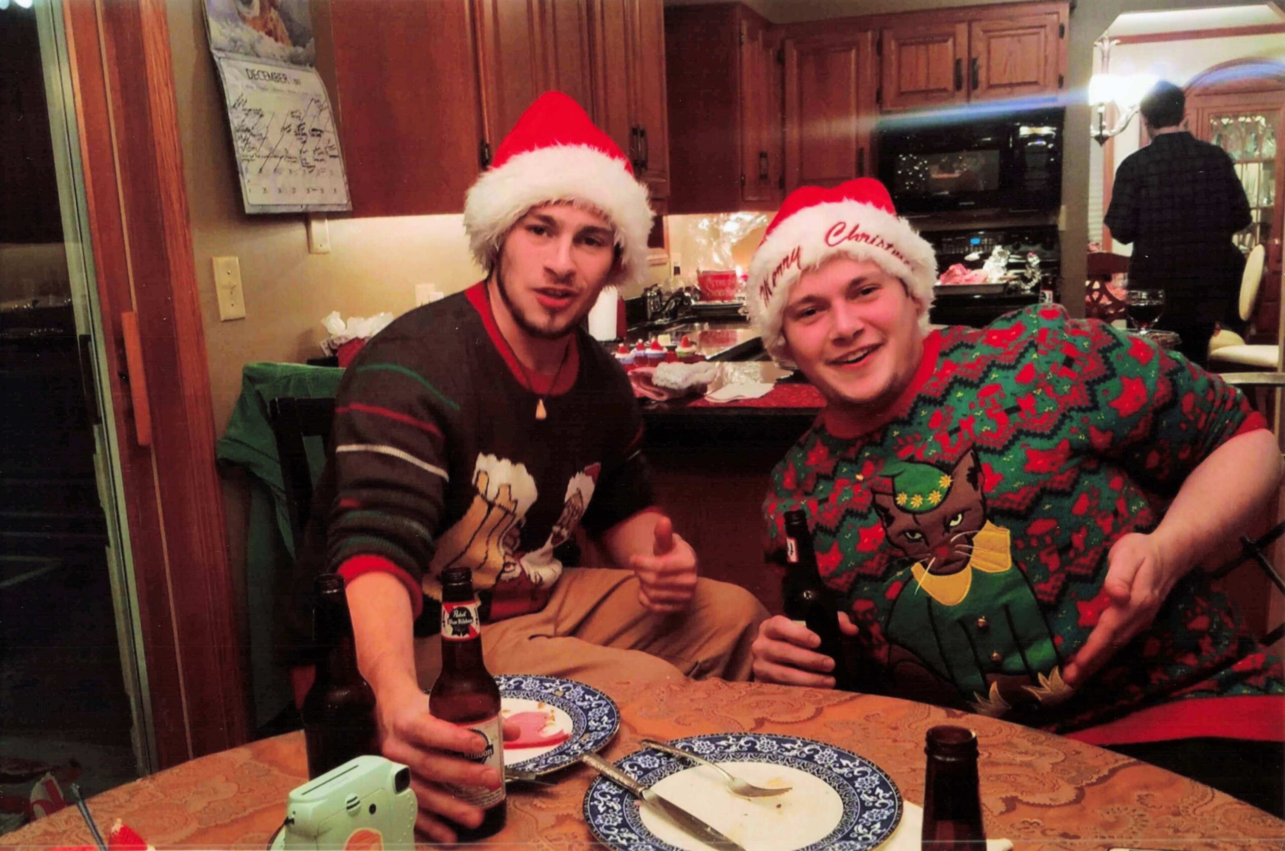 Two men wearing Santa hats sit at a dining table with blue-patterned plates and beer bottles, smiling at the camera.