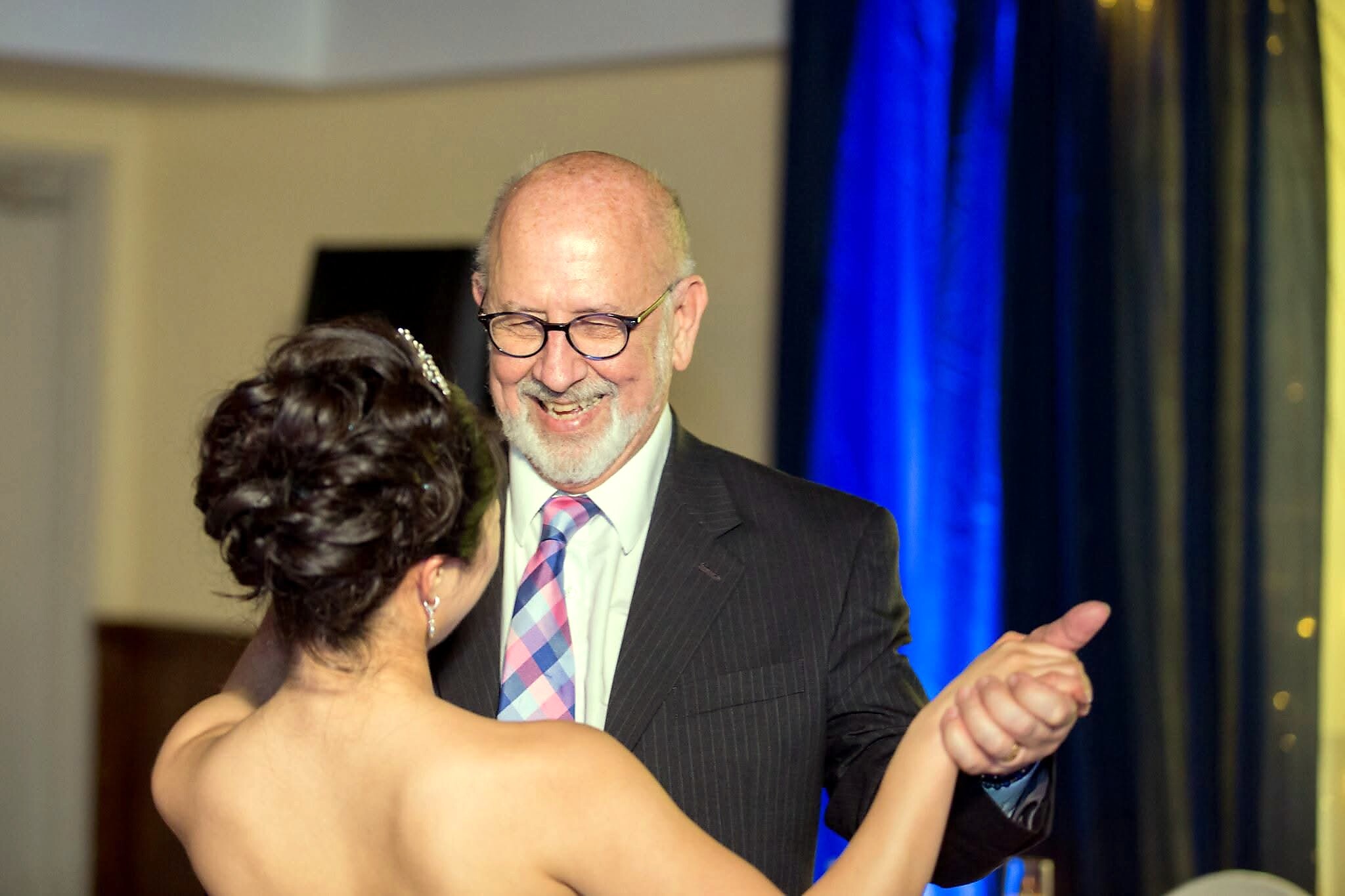 A smiling older man in a suit and tie dancing with a woman at a formal event, hands joined in a ballroom hold.