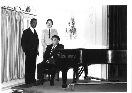 Three men in suits pose beside a Steinway grand piano in a formal room with a chandelier and heavy curtains, one seated at the piano.
