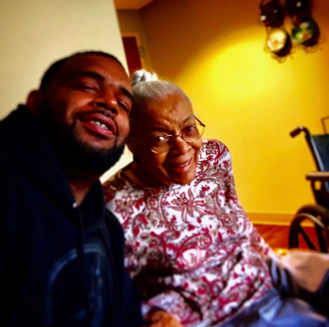 Smiling man and elderly woman taking a selfie indoors, yellow wall and a wheelchair visible in the background.