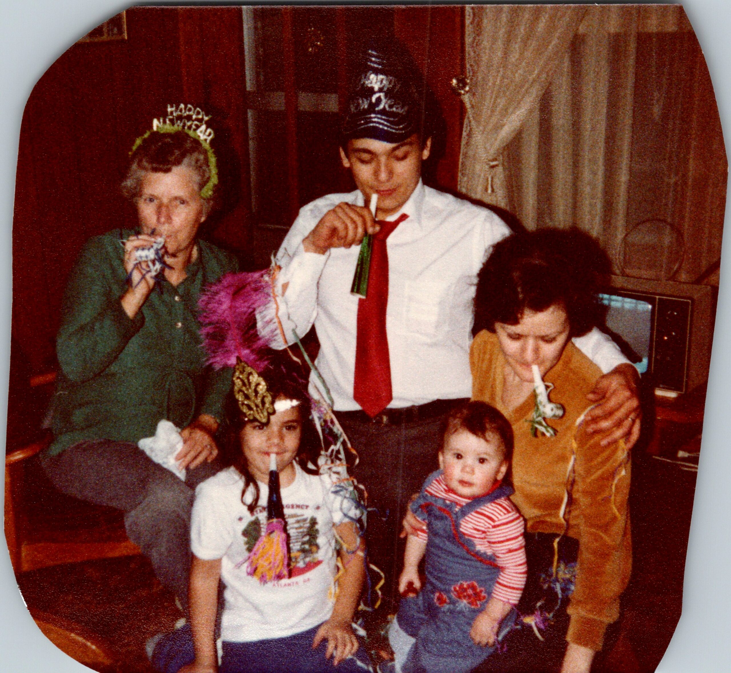 People celebrate New Year's Eve with party hats and decorations; a man in a white shirt and red tie holds a streamer while others drink and mingle nearby