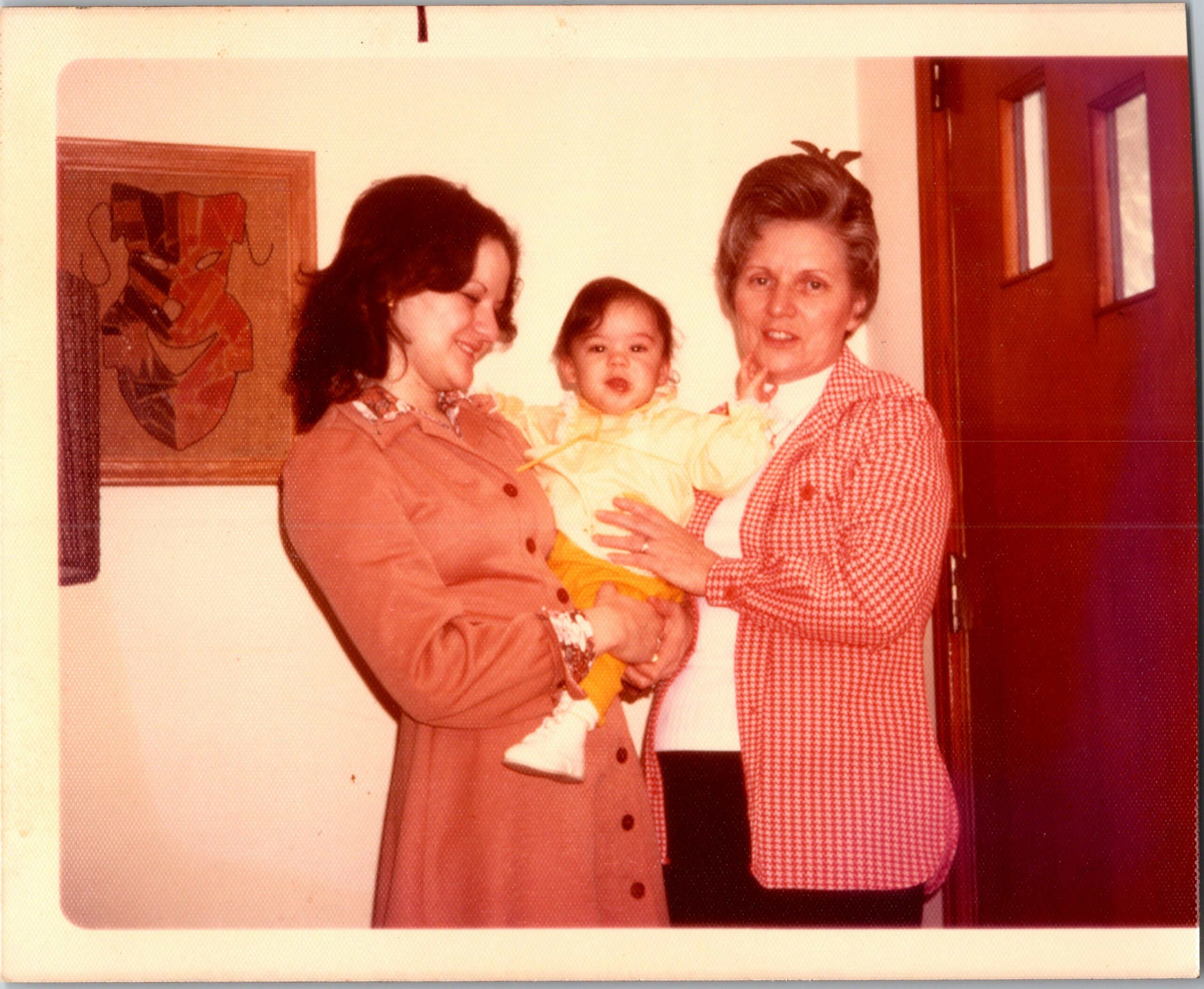 Two women hold a smiling baby indoors; one in a pink coat, the other in a red checkered jacket.