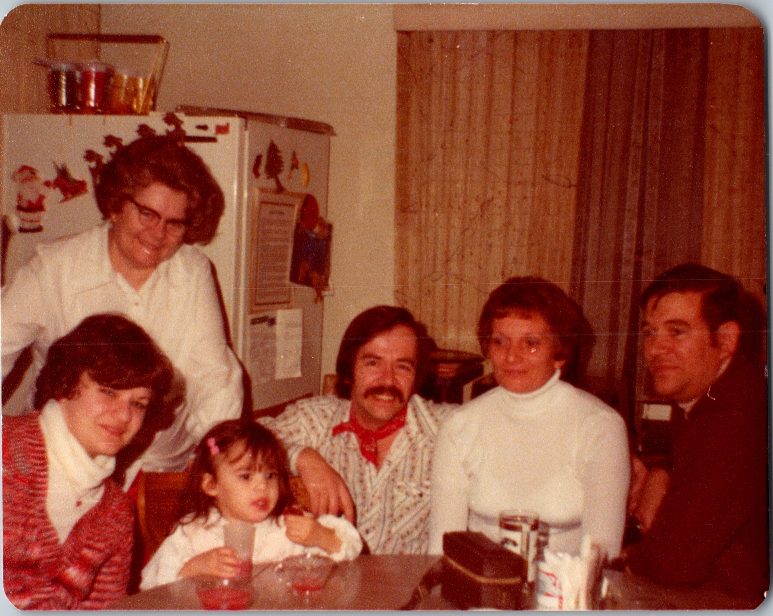 A smiling group of adults and children gathered in a home kitchen for a family photo, with a fridge in the background.