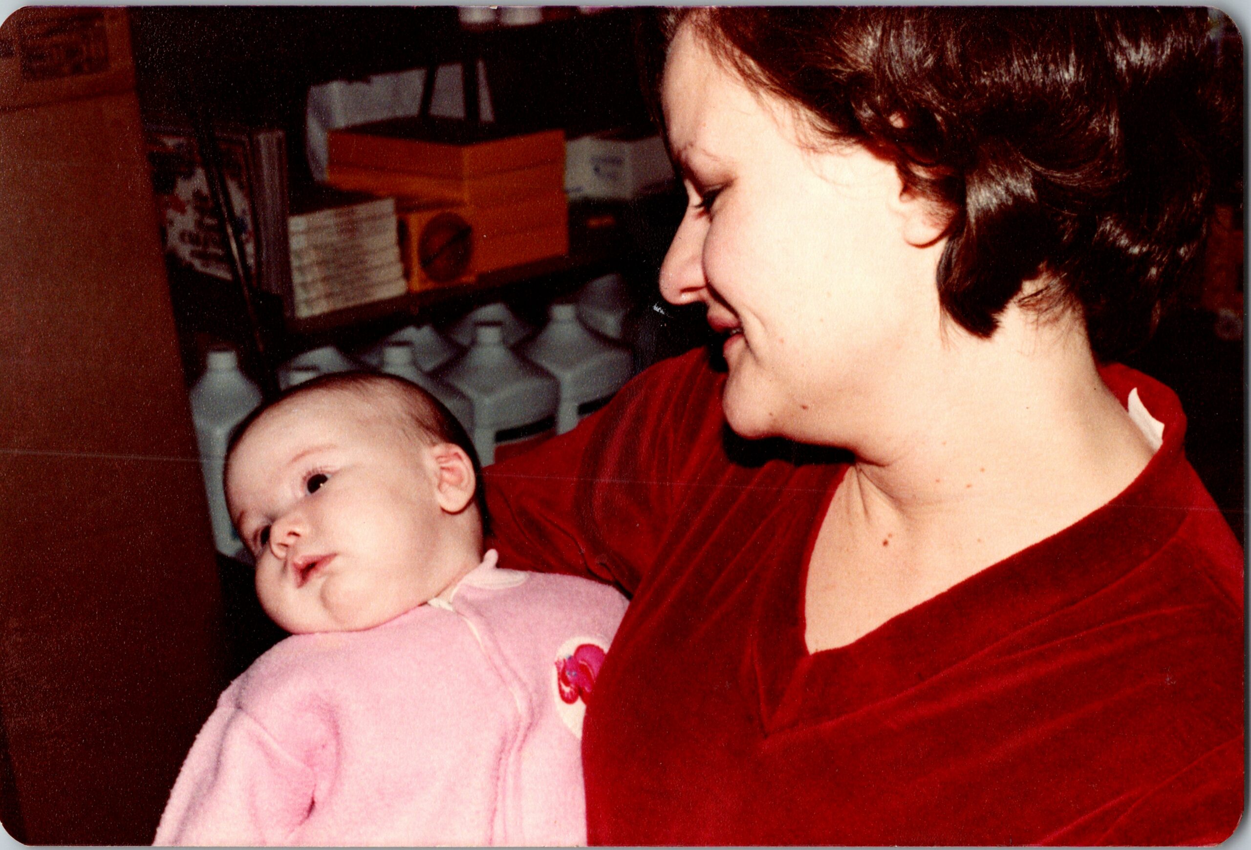 Woman in a red top holding a smiling infant in a pink outfit, both facing left.