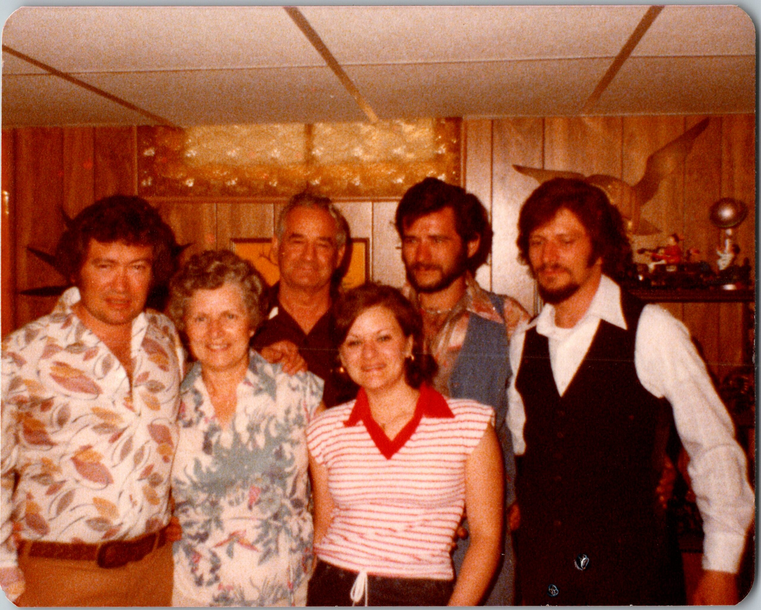 Group of people posing for a casual photo in a wood-paneled room, wearing 1970s-style clothing and smiles.