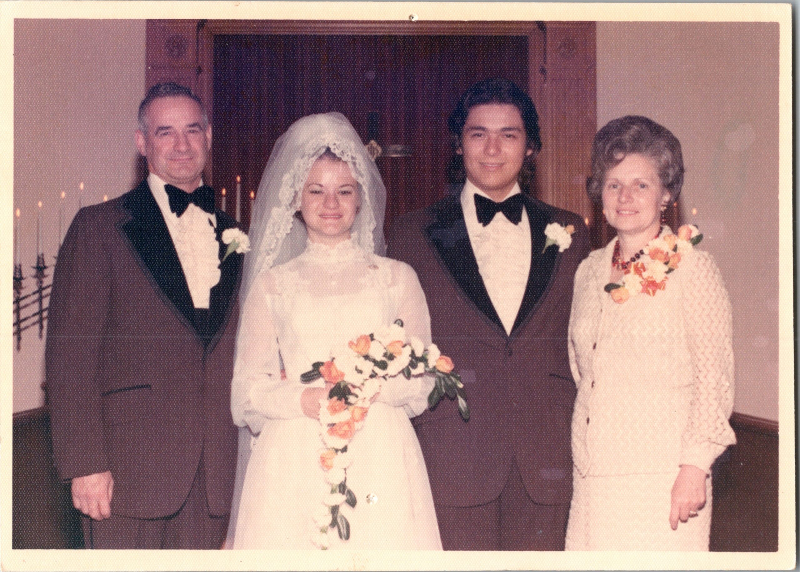 Wedding party portrait: bride in a white gown and veil with bouquet, flanked by a groom in a tuxedo and two guests indoors.