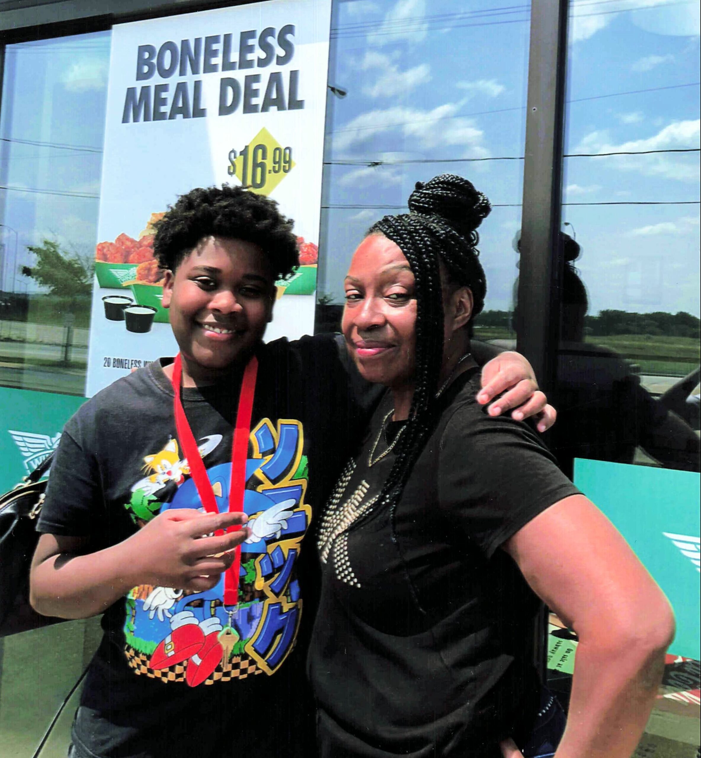 Two women smiling while posing for a selfie in front of a storefront banner that reads 'BONELESS MEAL DEAL'.