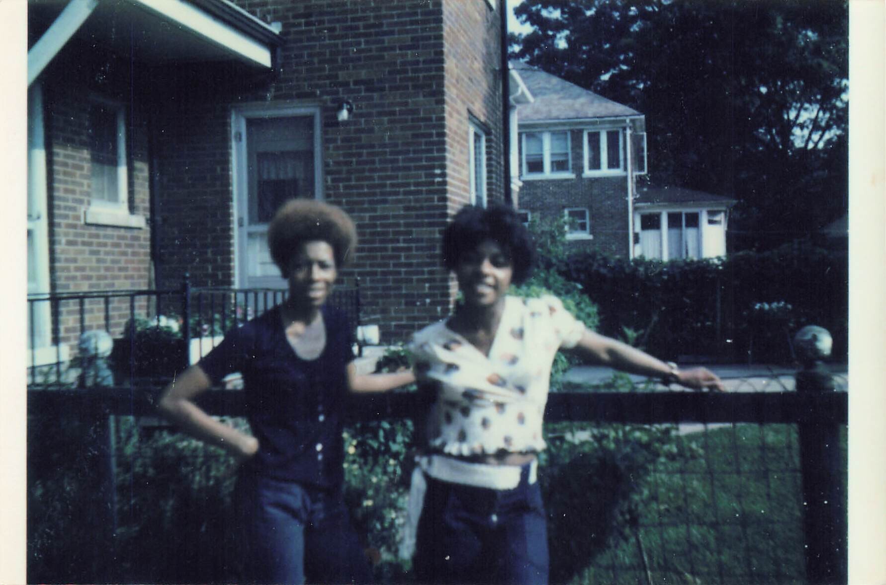 Two friends with afro hairstyles pose by a black iron fence in a suburban street, smiling at the camera.