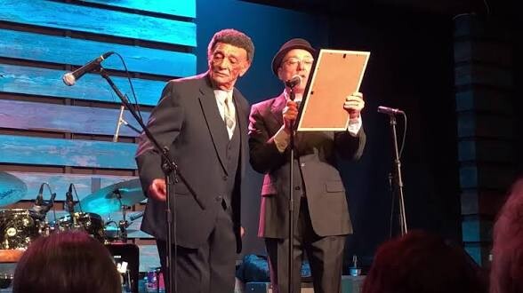 Two men in suits on a stage, one holding a clipboard as they speak into microphones during a live performance or presentation.