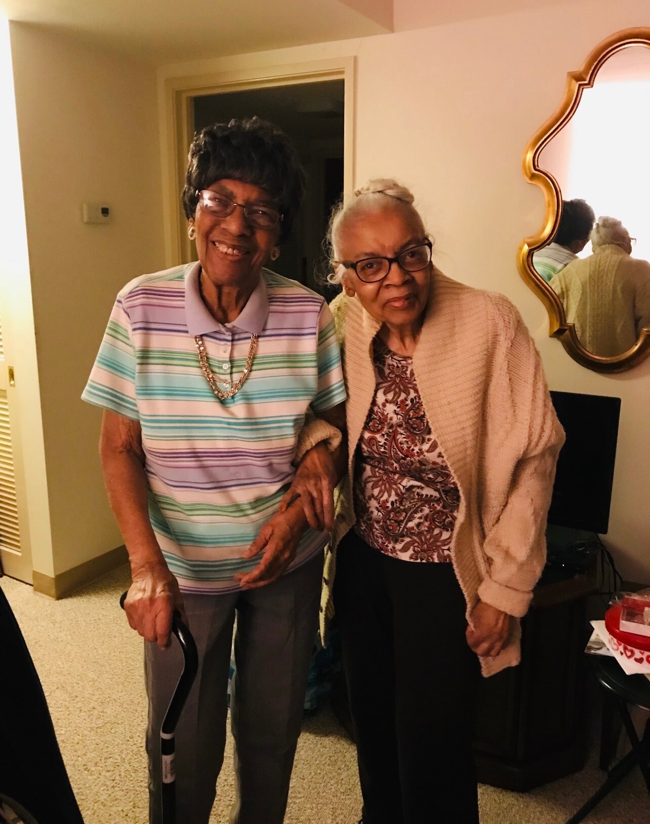 Two elderly women smiling and standing arm-in-arm in a living room.