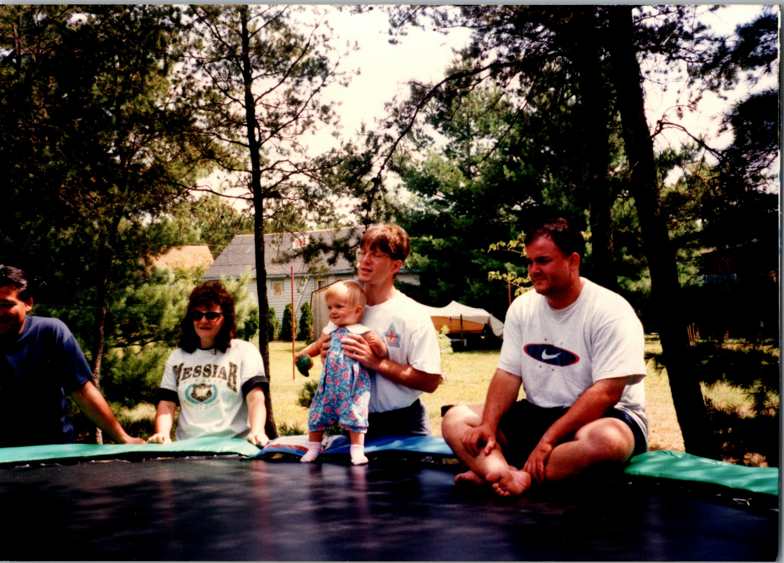 Family outdoors around a trampoline in a backyard, with adults and a small child standing on the mat, trees in the background.