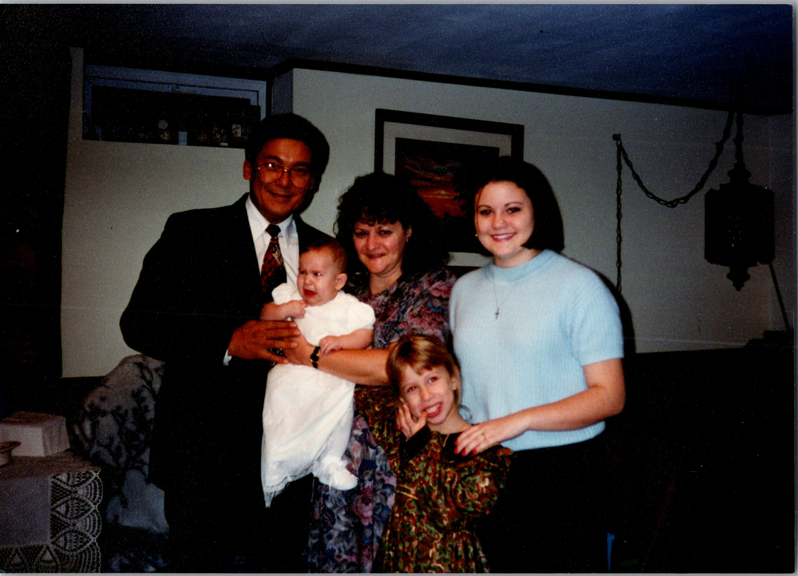 A family group in a living room: man in a suit holding a baby, flanked by two women and a smiling girl in a blue sweater.