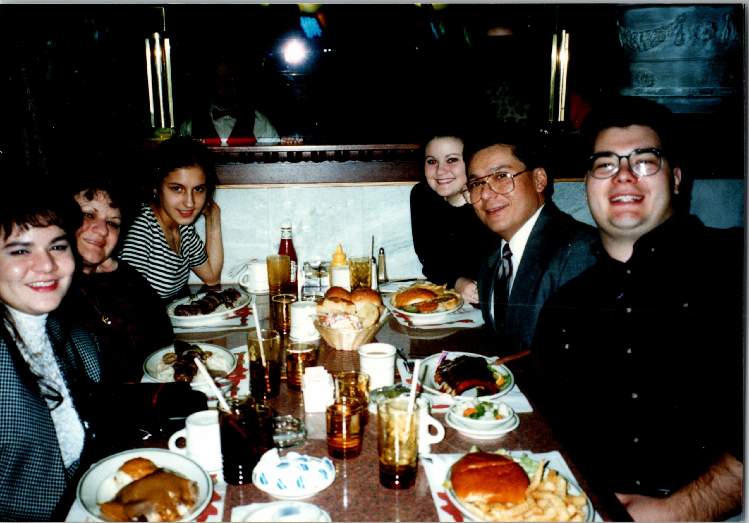 Group of friends dining at a restaurant table, smiling at the camera with burgers, fries, and drinks in view.