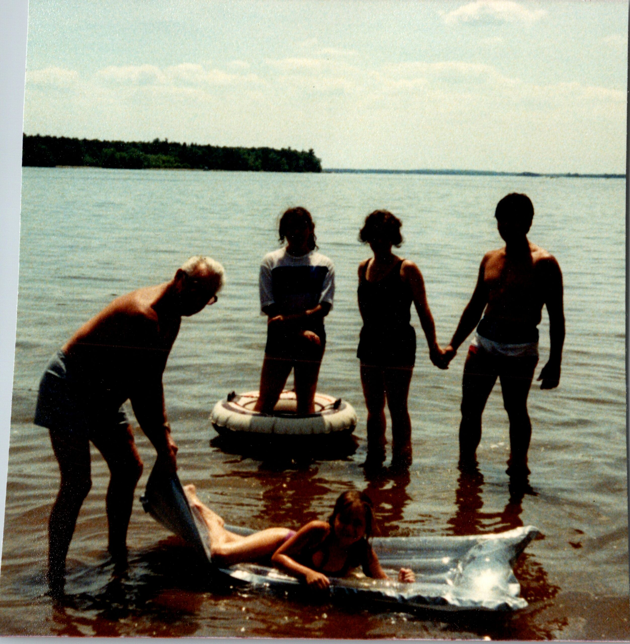Group of people standing in shallow water at the beach, with a white inflatable ring nearby and a tree-covered island in the distance.