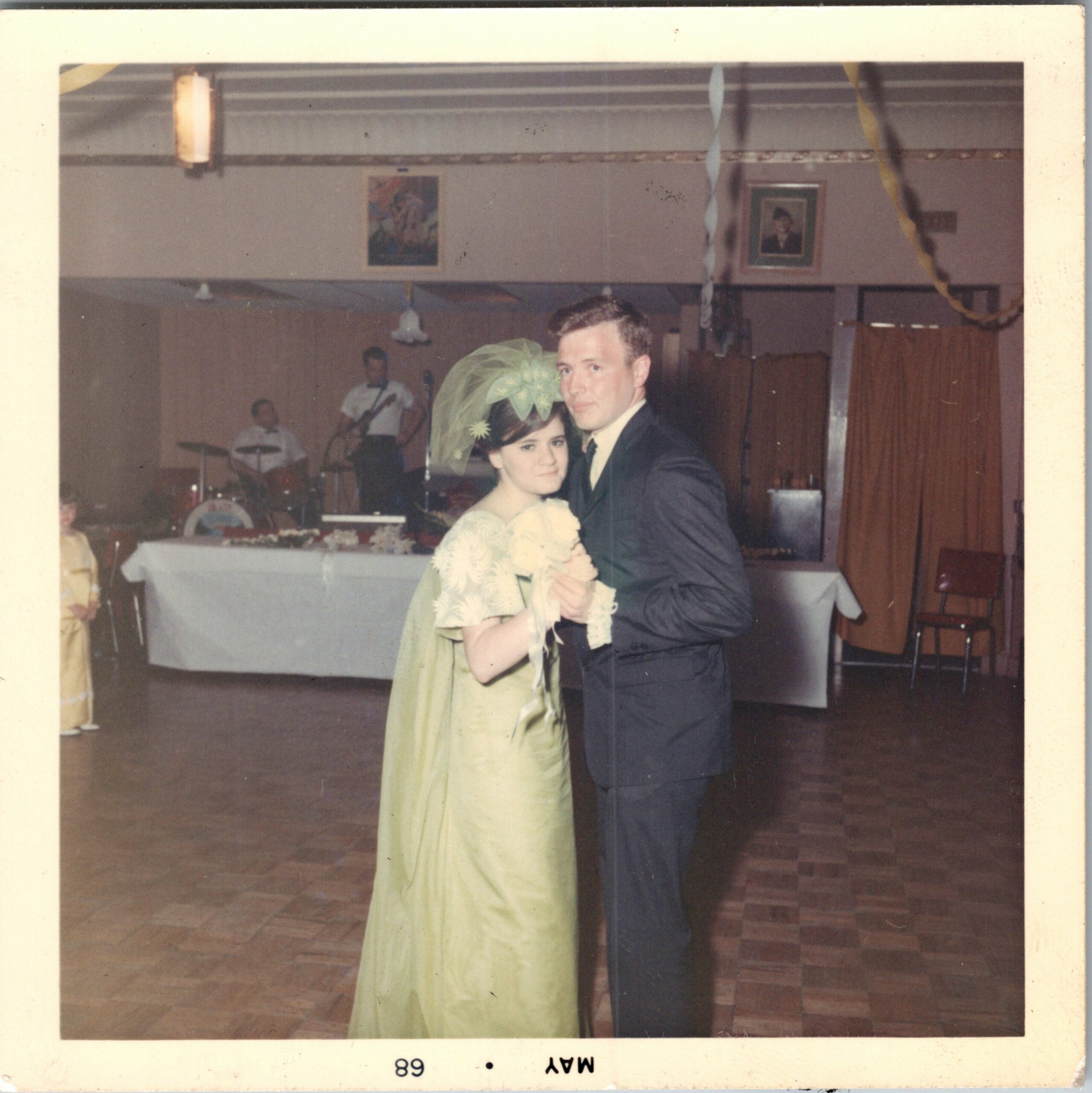 Couple dancing at an indoor celebration with streamers, a band in the background, and a woman in a light dress with a large floral headpiece nearby.