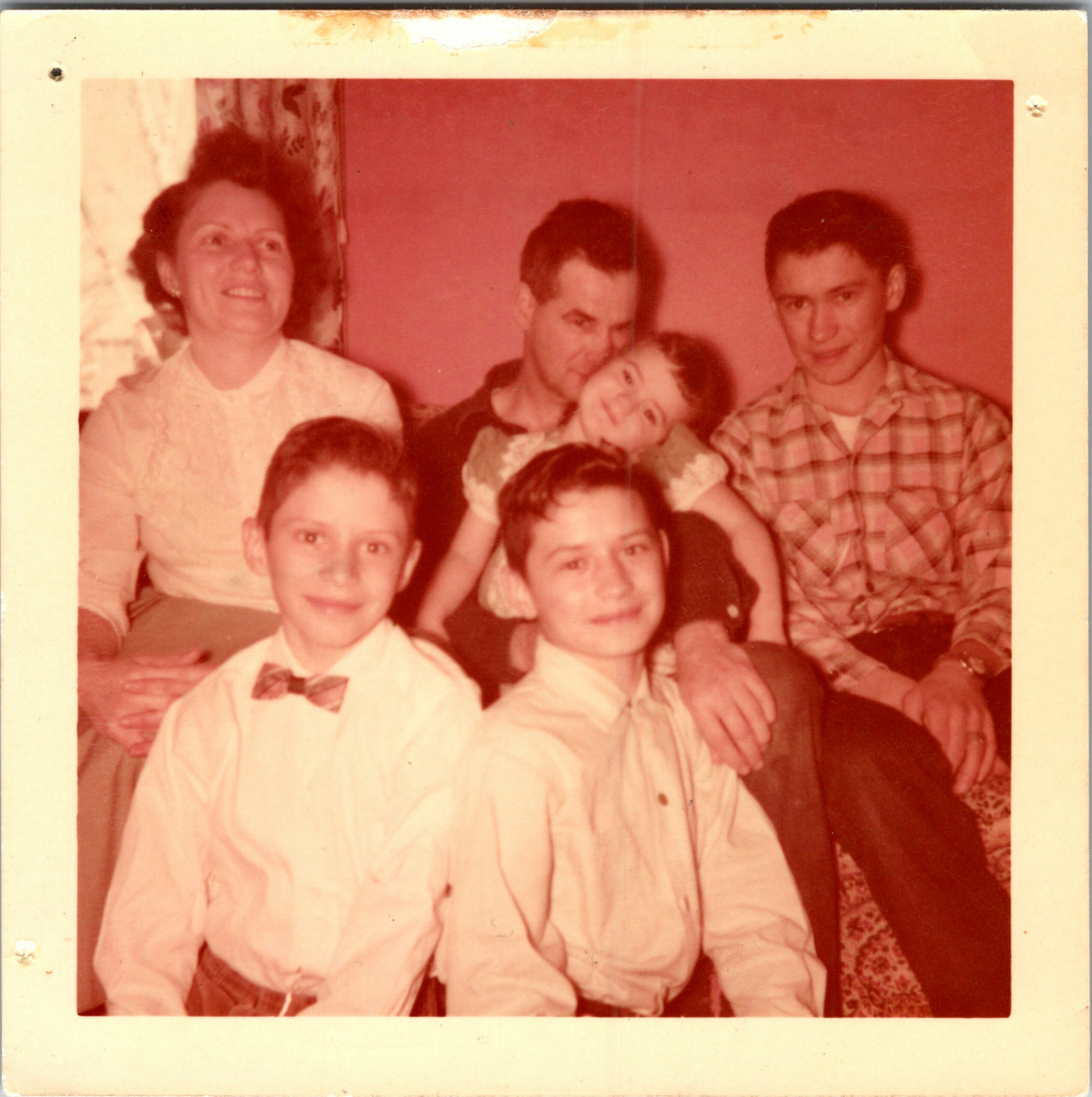 Group portrait of several people smiling and posing closely together in a warm, reddish-toned photo; appears to be a family or friends gathering indoors.