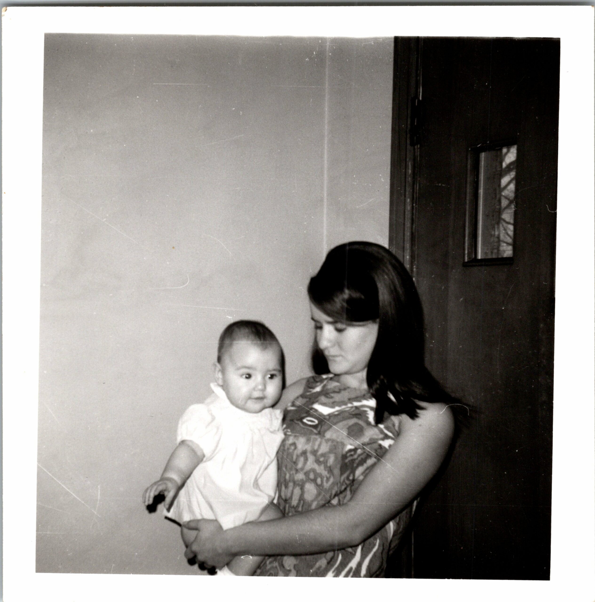 Mother cradling a baby in a white dress while standing indoors near a dark door and window.