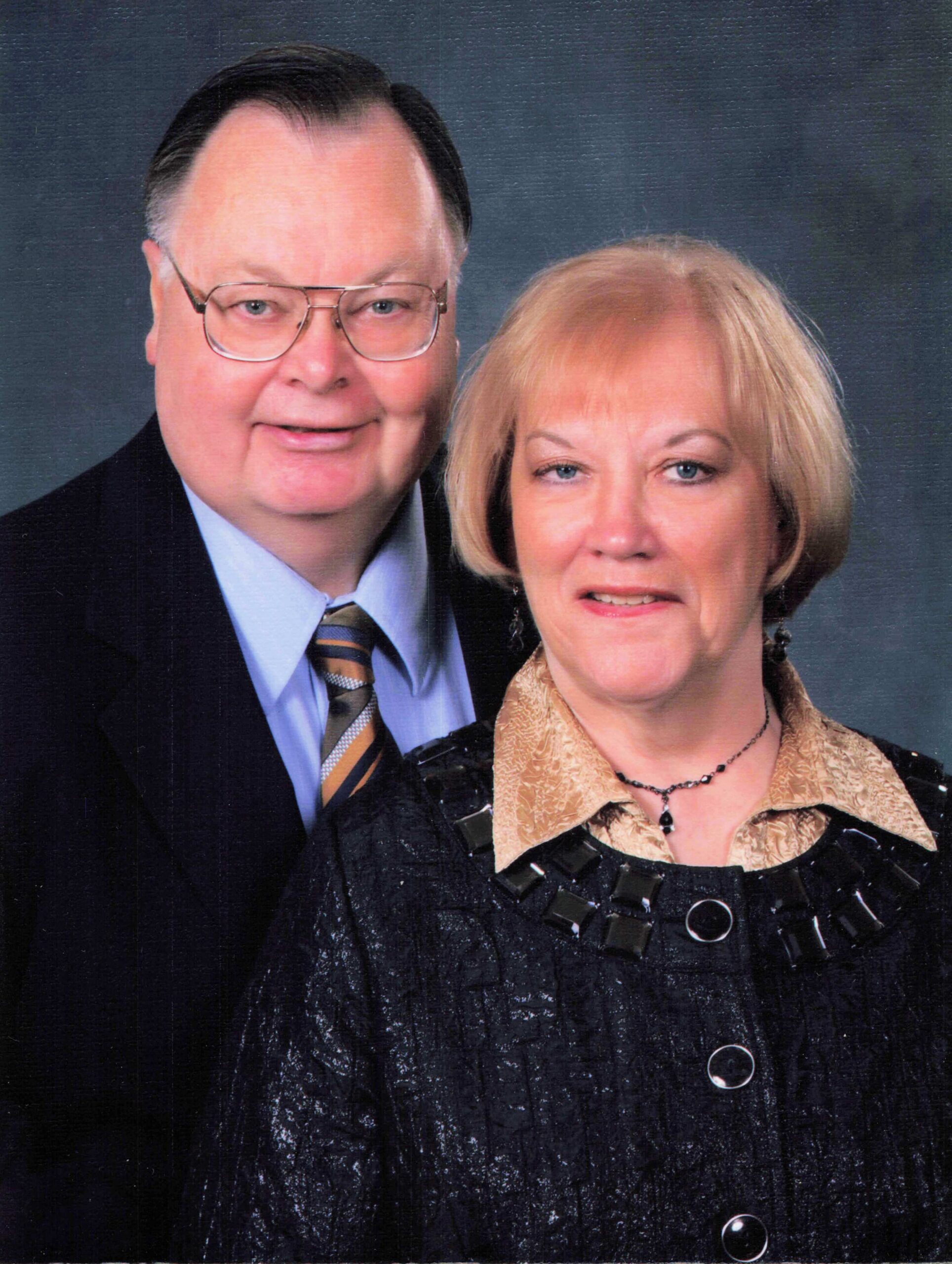 Two middle-aged adults posing for a studio portrait; man in a suit and striped tie, woman in a patterned blouse with necklace, against a dark backdrop.