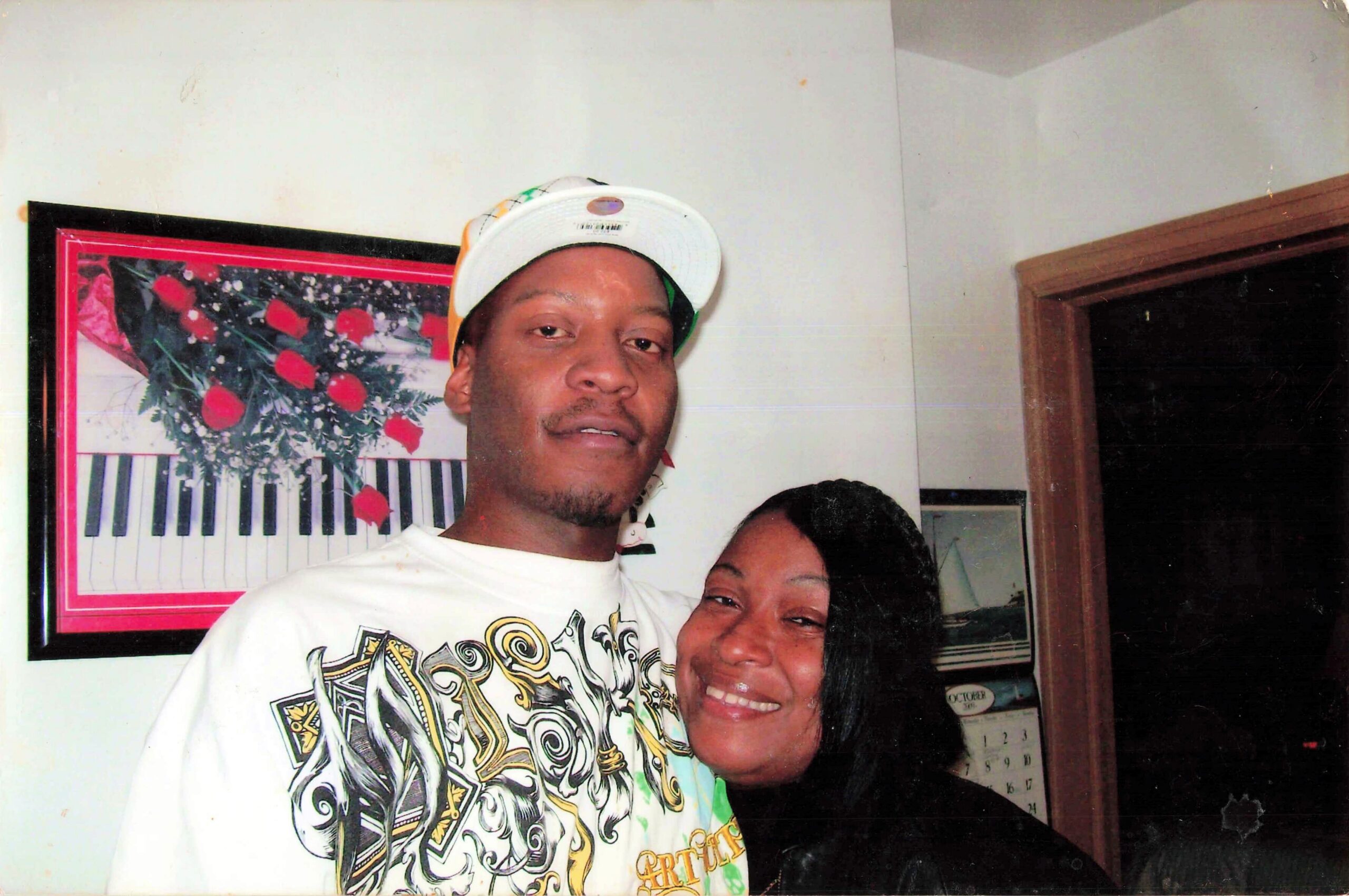Two friends posing for a casual indoor photo; man in a white cap and graphic shirt stands beside a smiling woman indoors by a framed artwork and calendar.