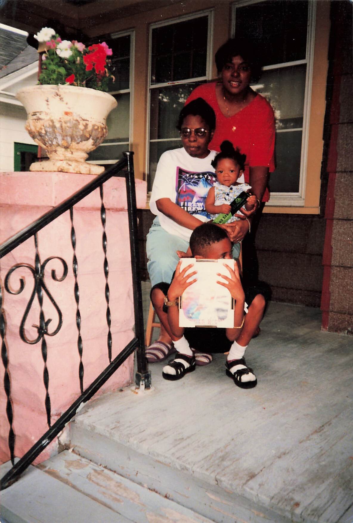 Three people on a front porch: a seated woman with glasses holding a small child, a second woman in red standing behind them, and a child sitting on the steps with a book/album