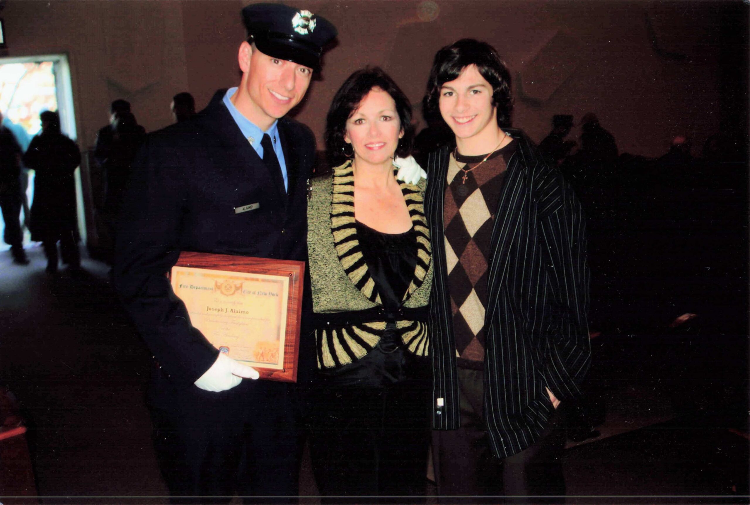 Three people posing together: a man in a police uniform holding a certificate, flanked by a woman in a striped gold outfit and a young man in a brown argyle sweater vest.