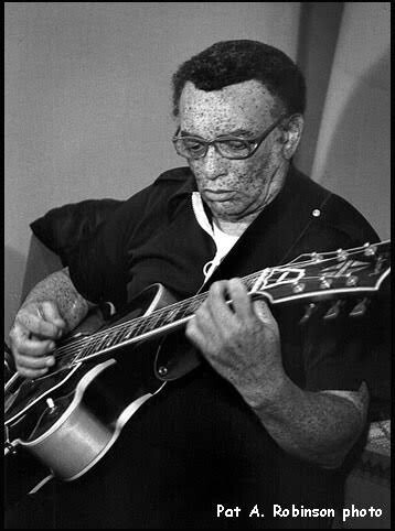 Man wearing glasses plays an acoustic guitar, seated and focused on the strings in a black-and-white photo.