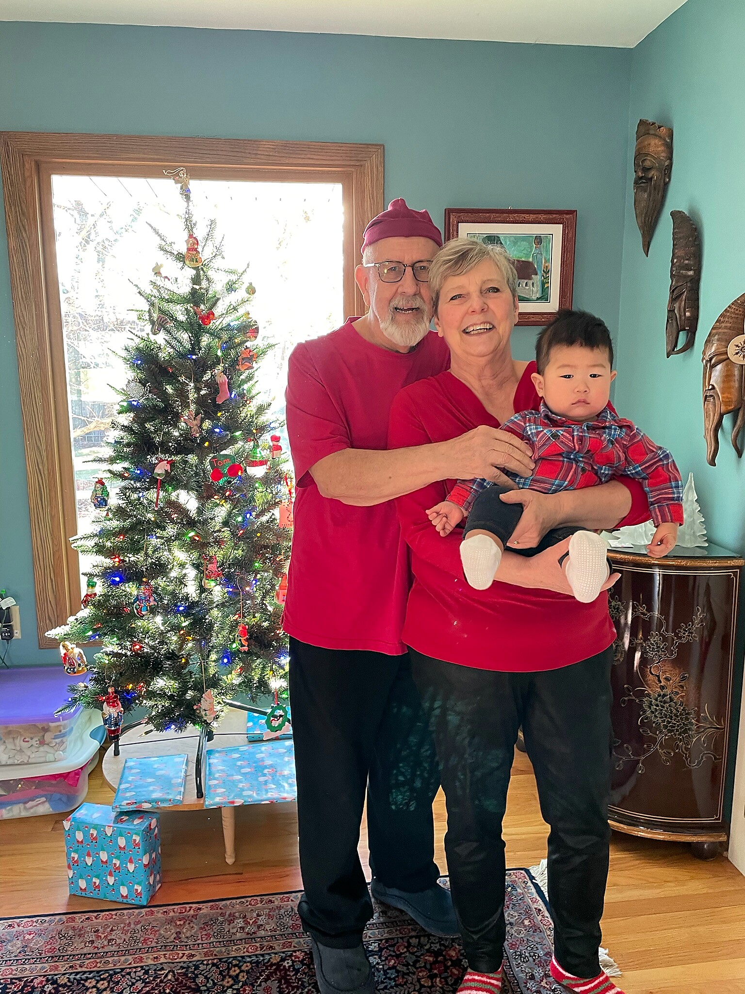 Smiling couple in red shirts hug a toddler by a decorated Christmas tree in a bright living room.