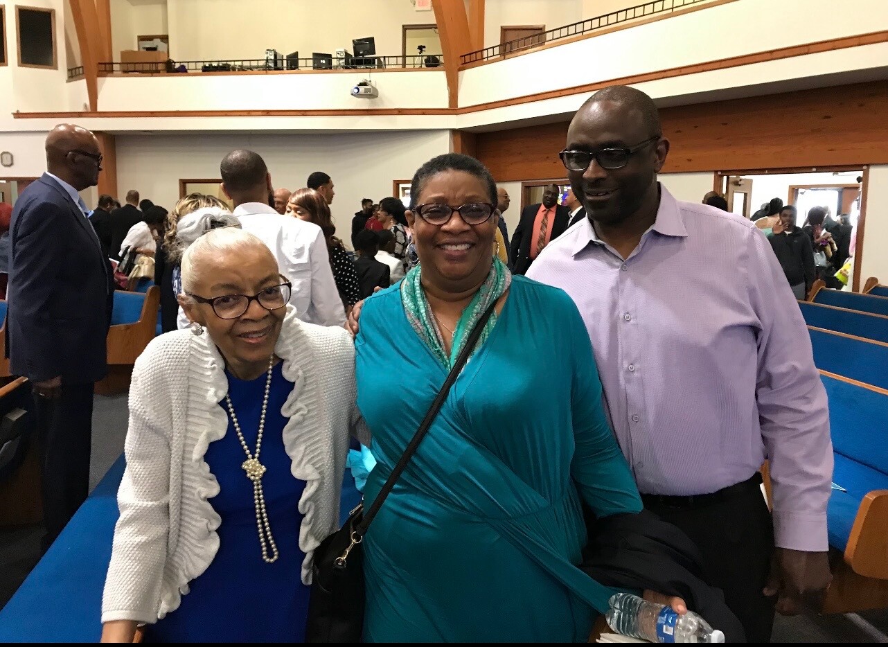 Three adults pose for a photo in a church hall, smiling at the camera: an elderly woman in a white cardigan and blue dress, a woman in a teal dress with scarf, and a man in a lavender shirt beside them; she holds a water bottle.