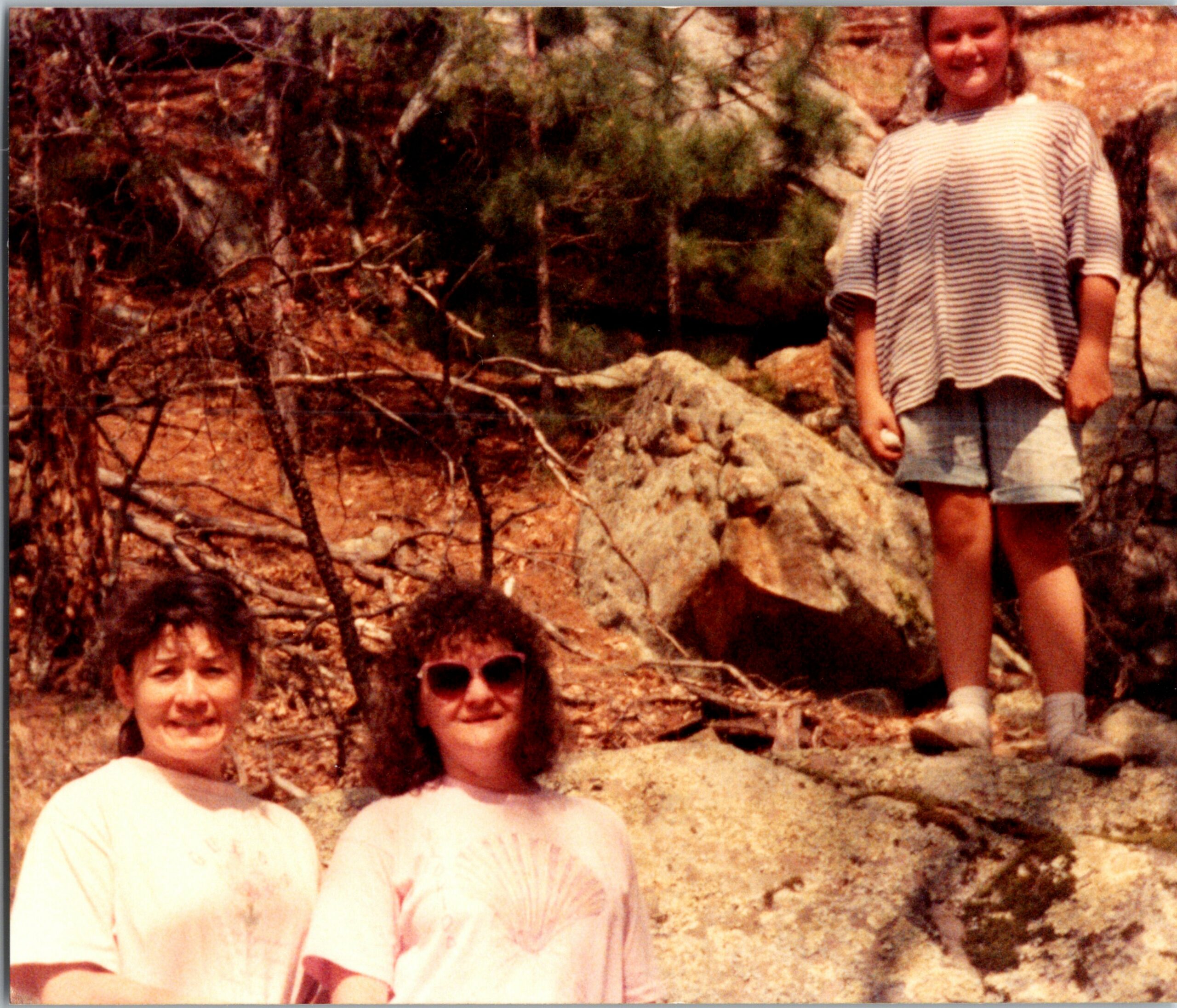 Group of children posing outdoors on a rocky hillside, partial view of two people standing and others seated below, wearing casual clothing.
