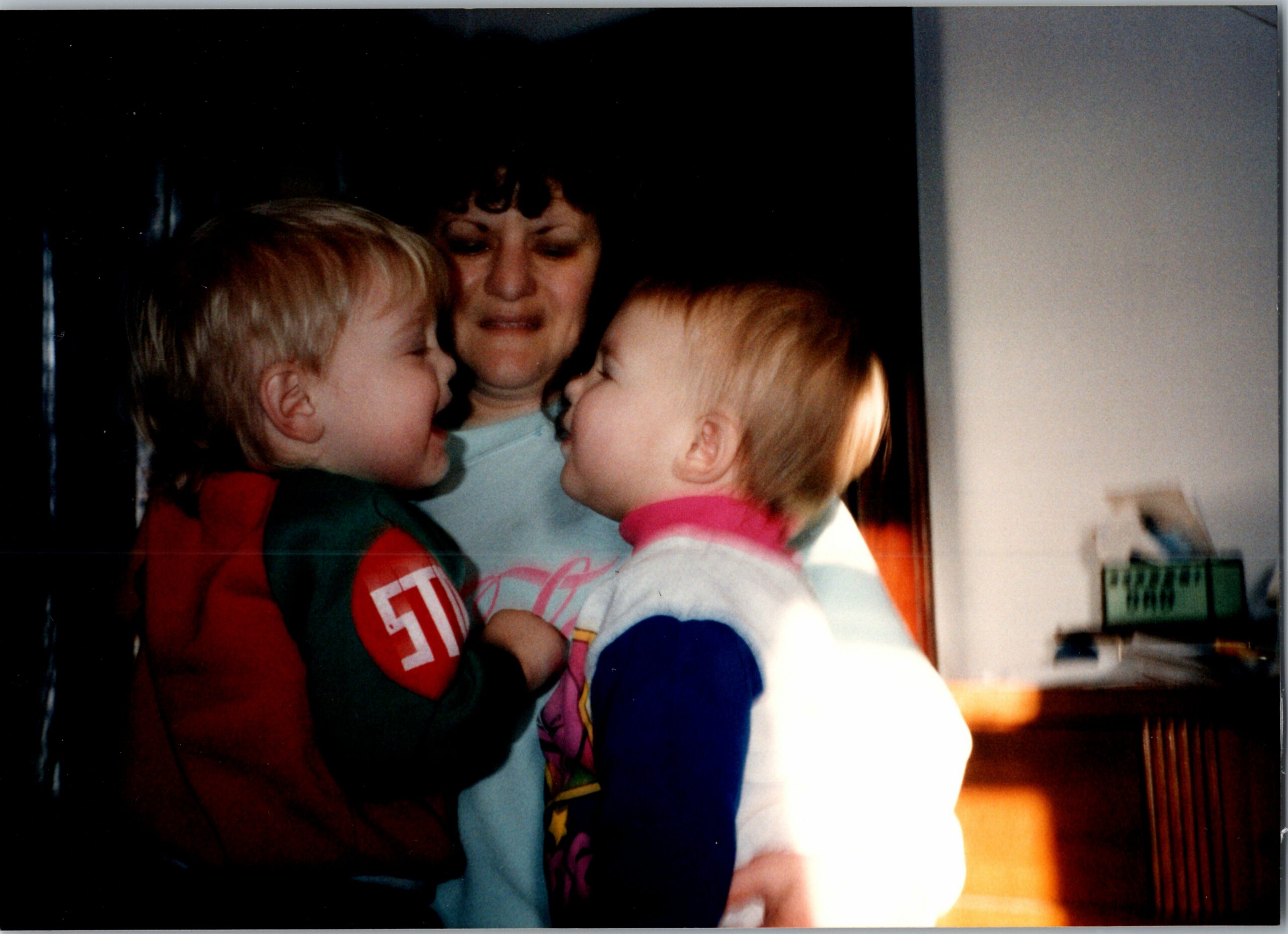 A woman holds two young children in a close hug, faces near each other in a warm family moment.