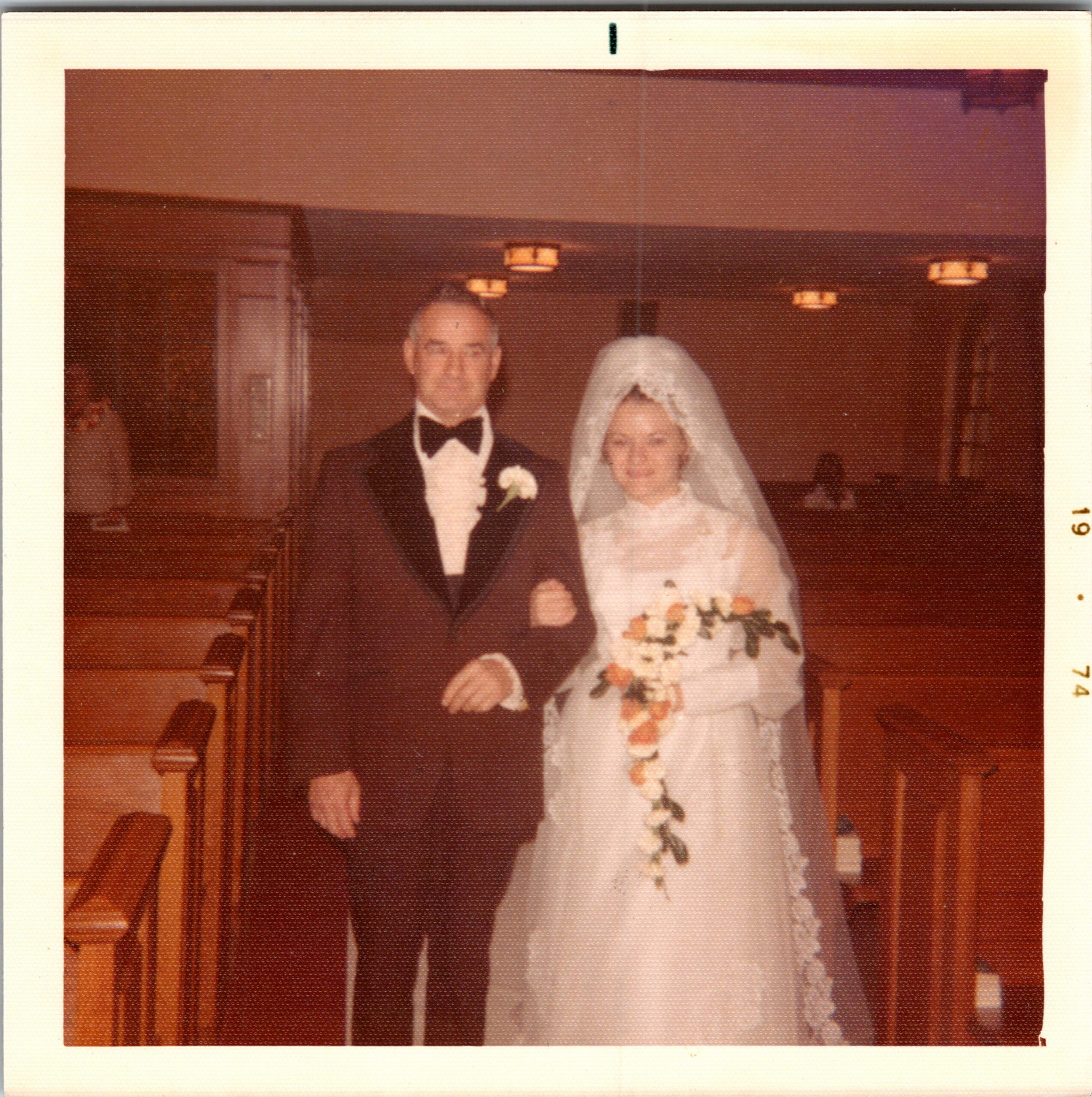 Bride in a white wedding dress with a veil stands next to a groom in a black tuxedo with a boutonniere; indoor wedding setting behind them.