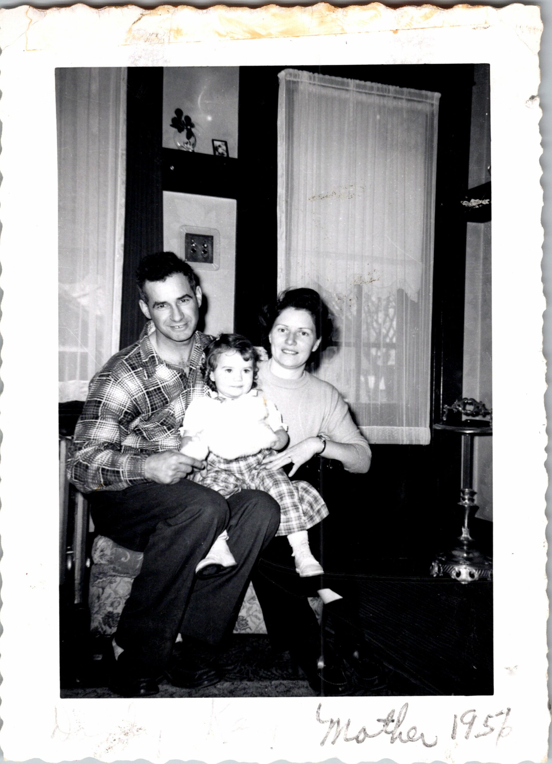 Family of three posing for a candid portrait in a living room, mother holding an infant while father sits beside them in a plaid shirt and dark pants.