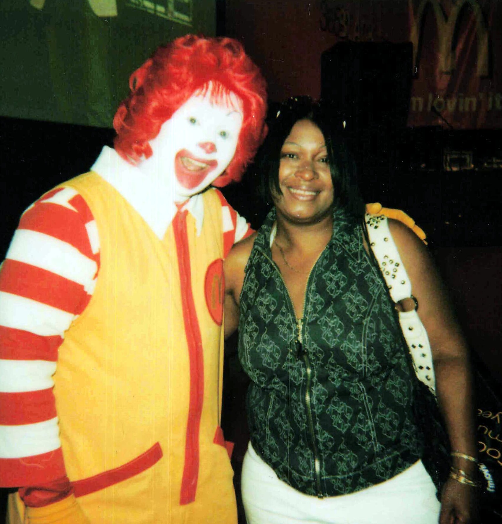 Clown with bright red hair and yellow costume posing with a smiling woman in a patterned sleeveless top.
